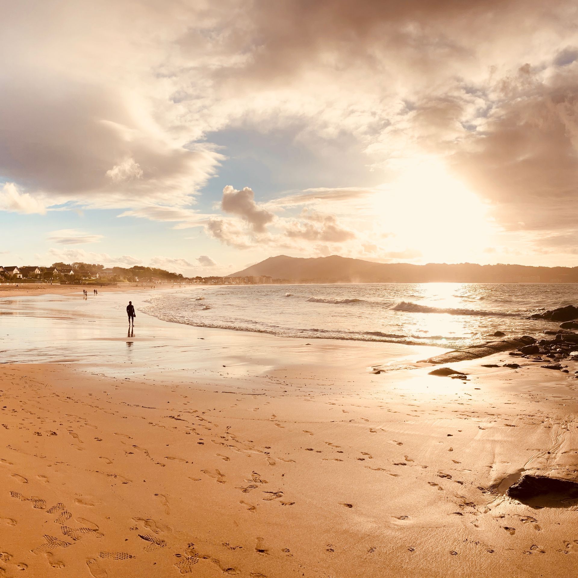 Person walking on beach at sunset; golden light, waves, and cloudy sky.