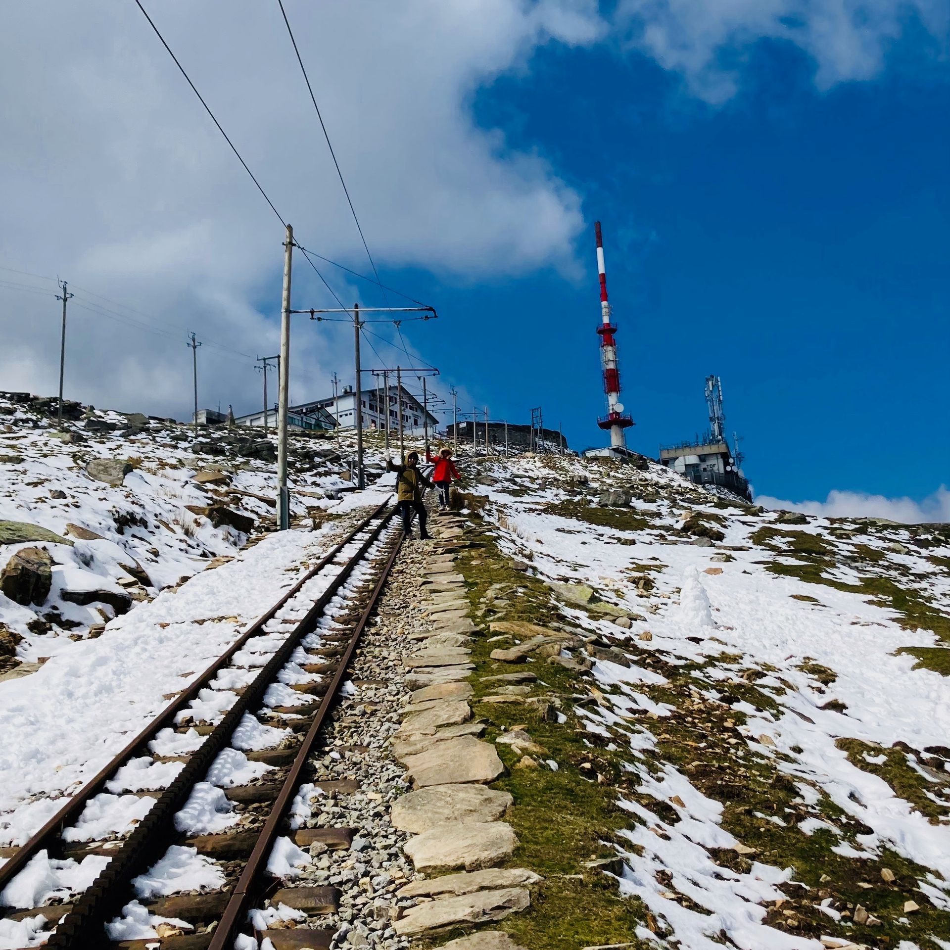 Railroad tracks ascend a snow-covered mountain. Red and white communication tower and buildings on the peak.