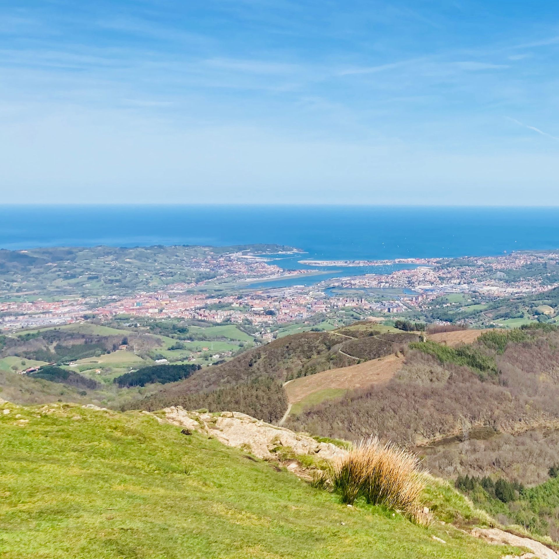View from mountain overlooking a coastal city, blue ocean, green fields, and clear sky.