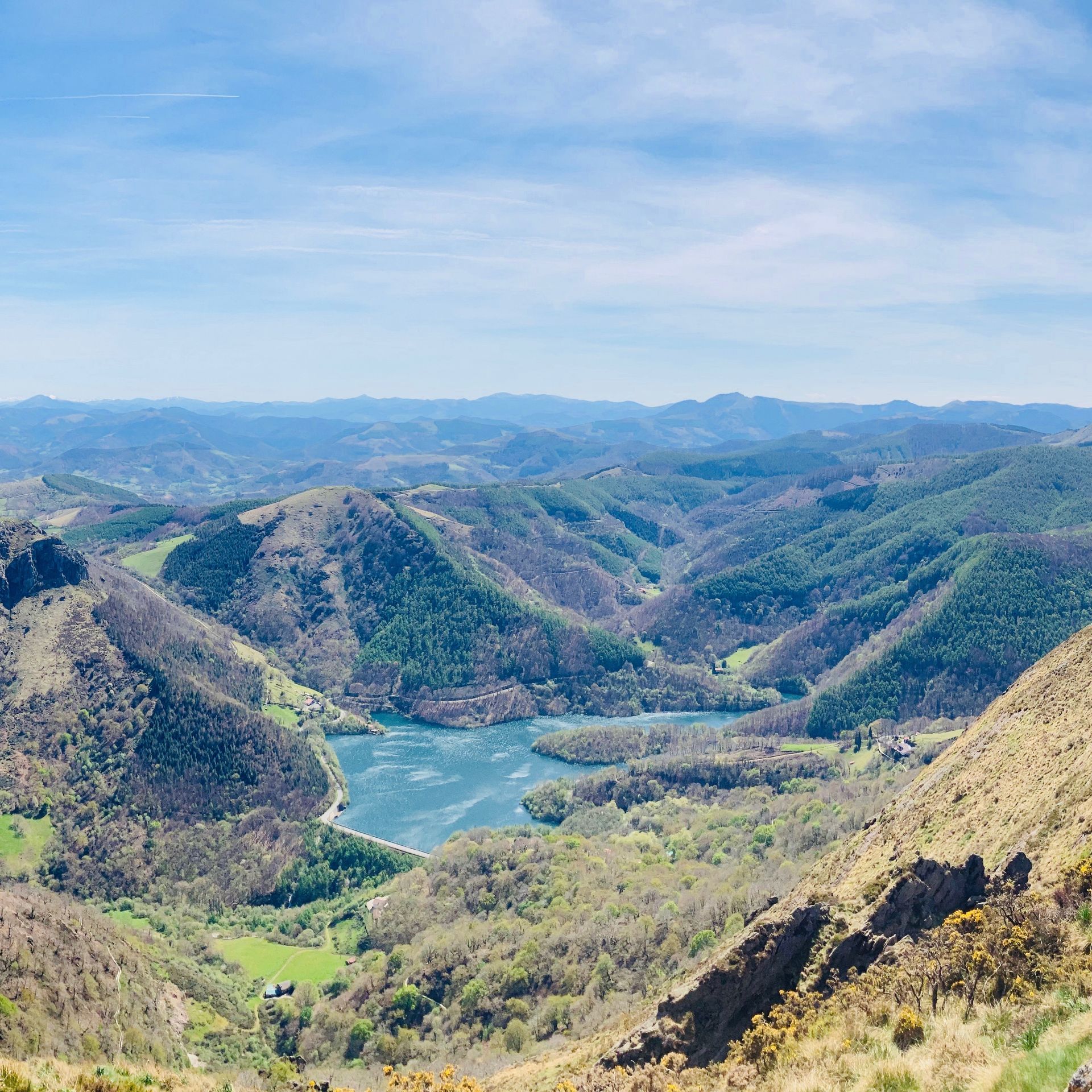 Panoramic view of a valley with a lake surrounded by green hills under a blue sky.