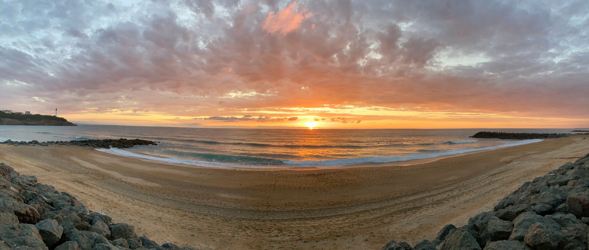 Sunset over a sandy beach and ocean with rocks on either side, orange sky and clouds.