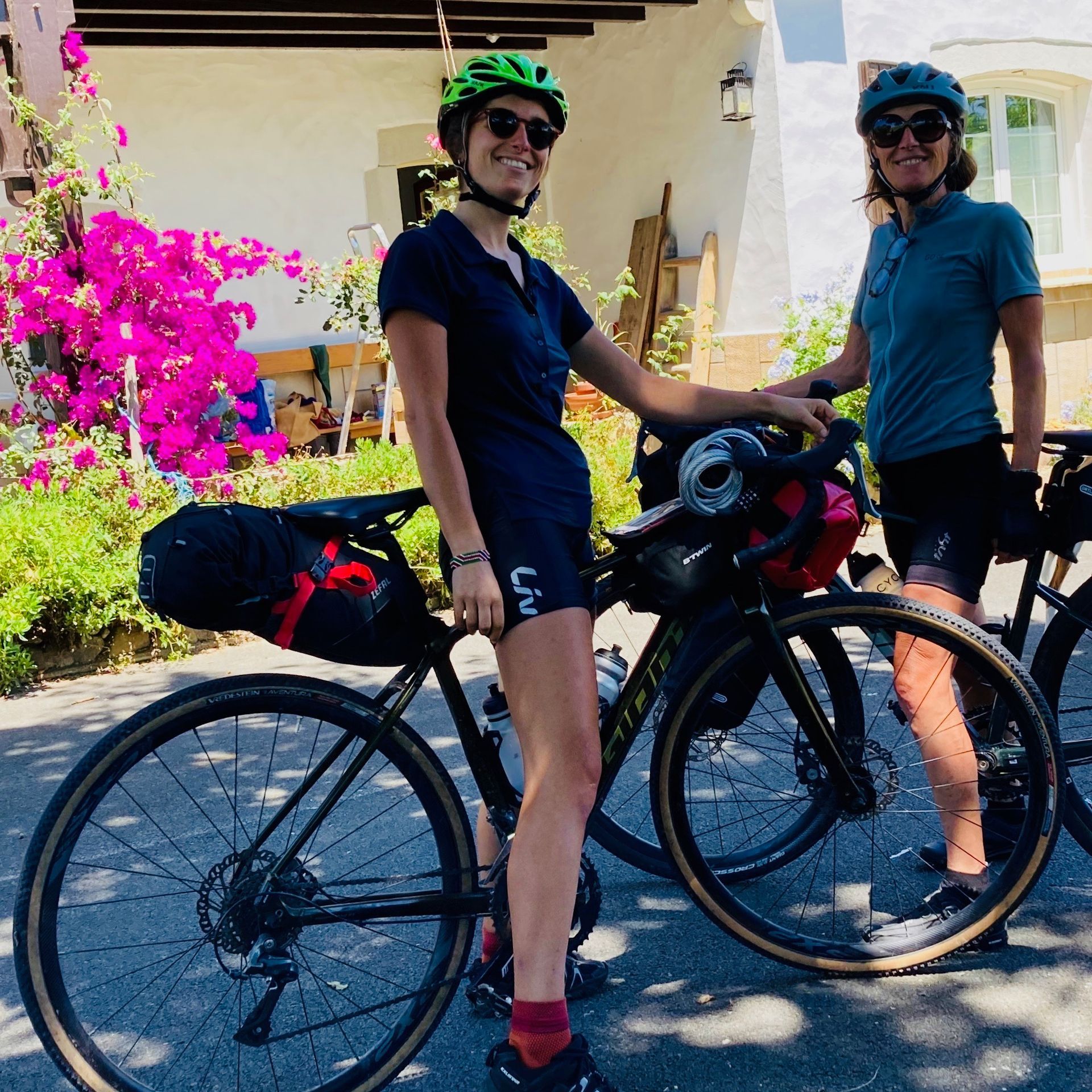Two women on bikes posing near a whitewashed building with pink flowers.