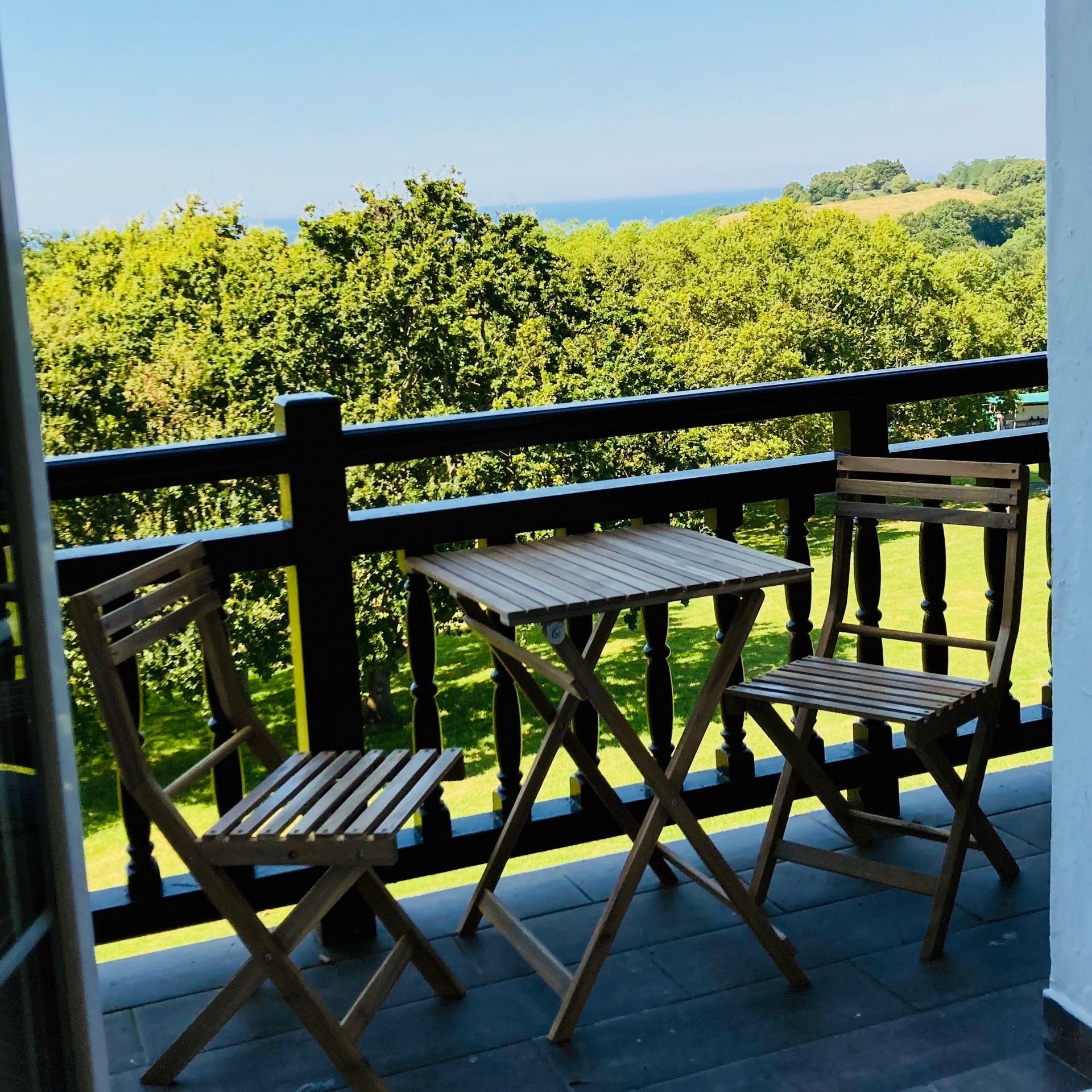 Balcony with wooden furniture, overlooking green trees and ocean under a blue sky.