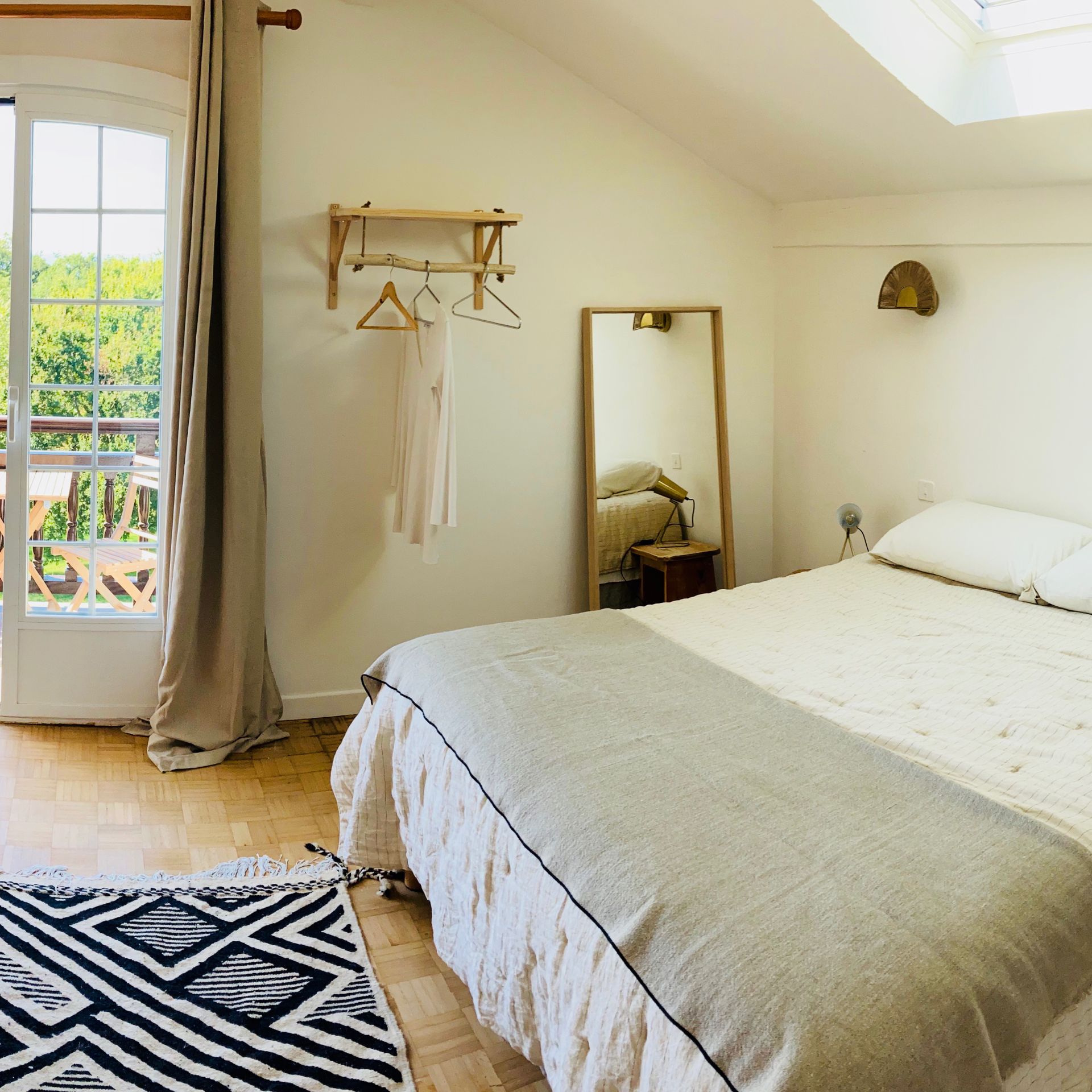 Bedroom with bed, full-length mirror, rug, open doorway to balcony. Soft, neutral tones; natural light.