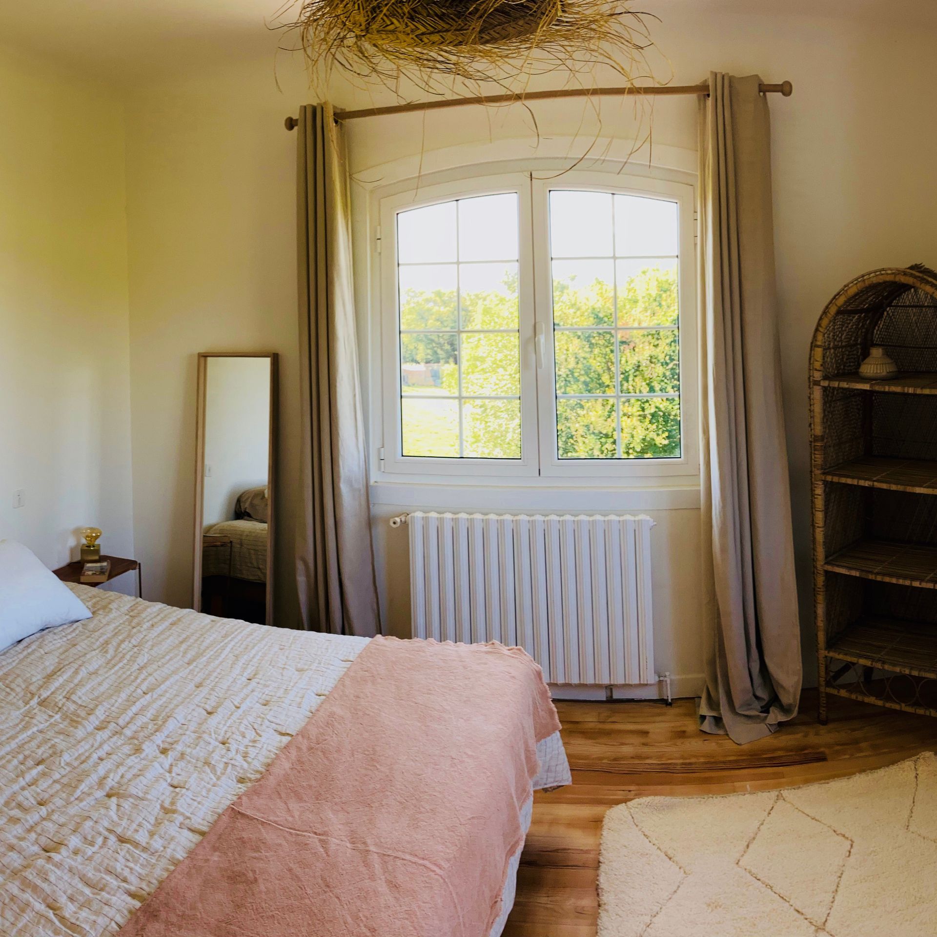 Bedroom with bed, window, mirror, and woven shelves. Soft pink and beige hues.