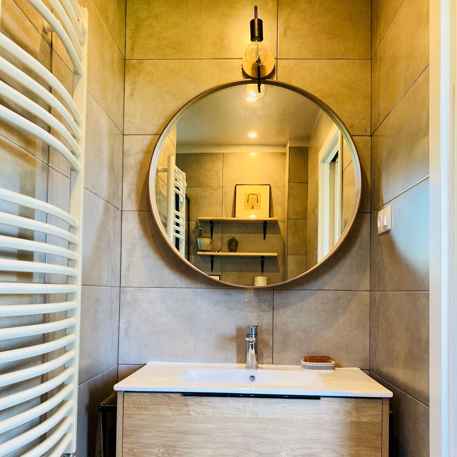 Bathroom with round mirror over a sink and wooden cabinet; light beige tiled walls; towel rack.