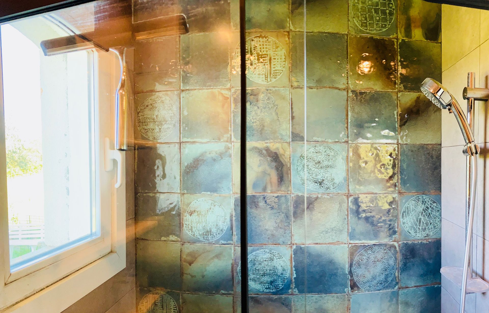 Shower with metallic green and blue square tiles, a window, and a showerhead.
