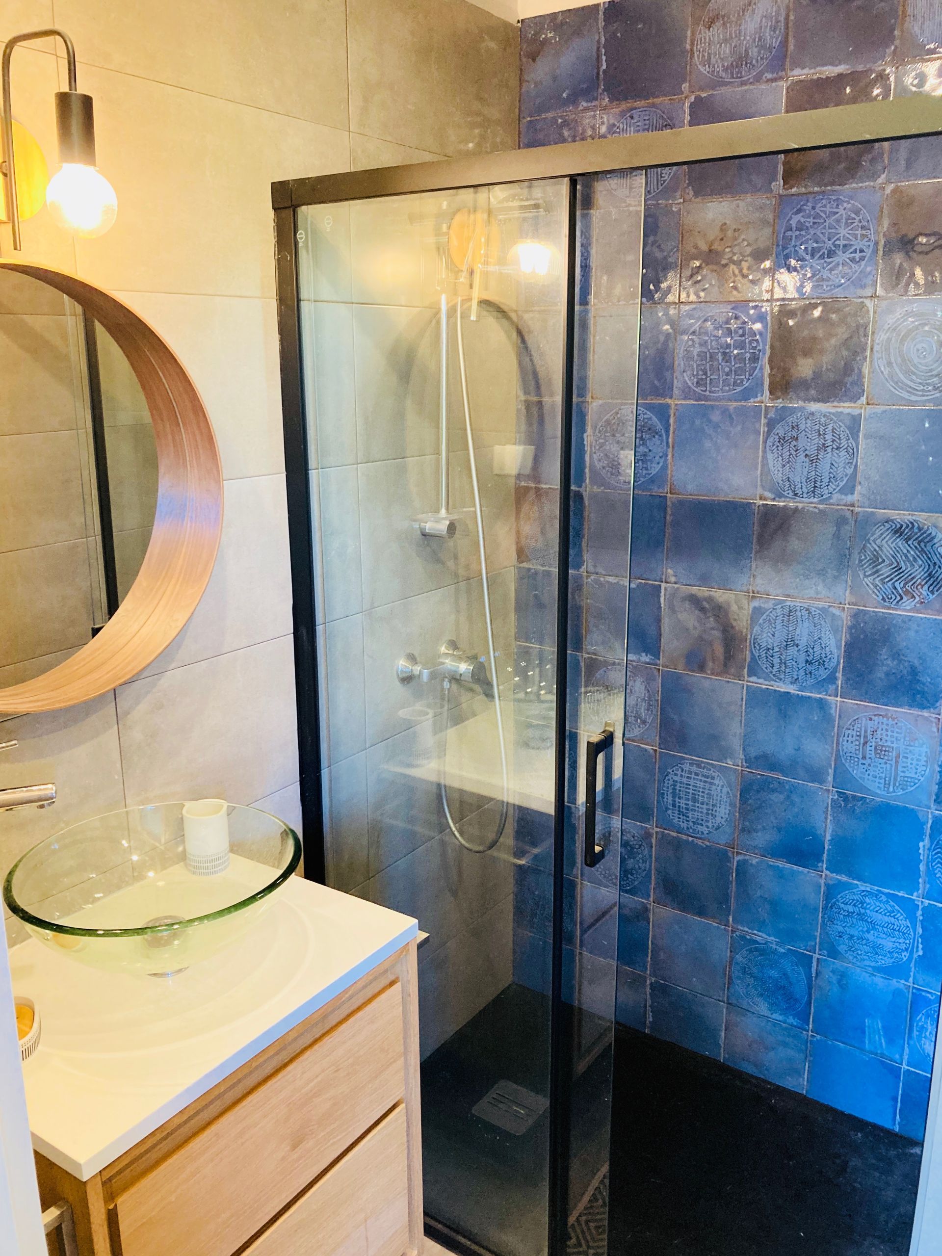 Bathroom with blue tiled shower, glass door, wooden vanity, round mirror, and sink.