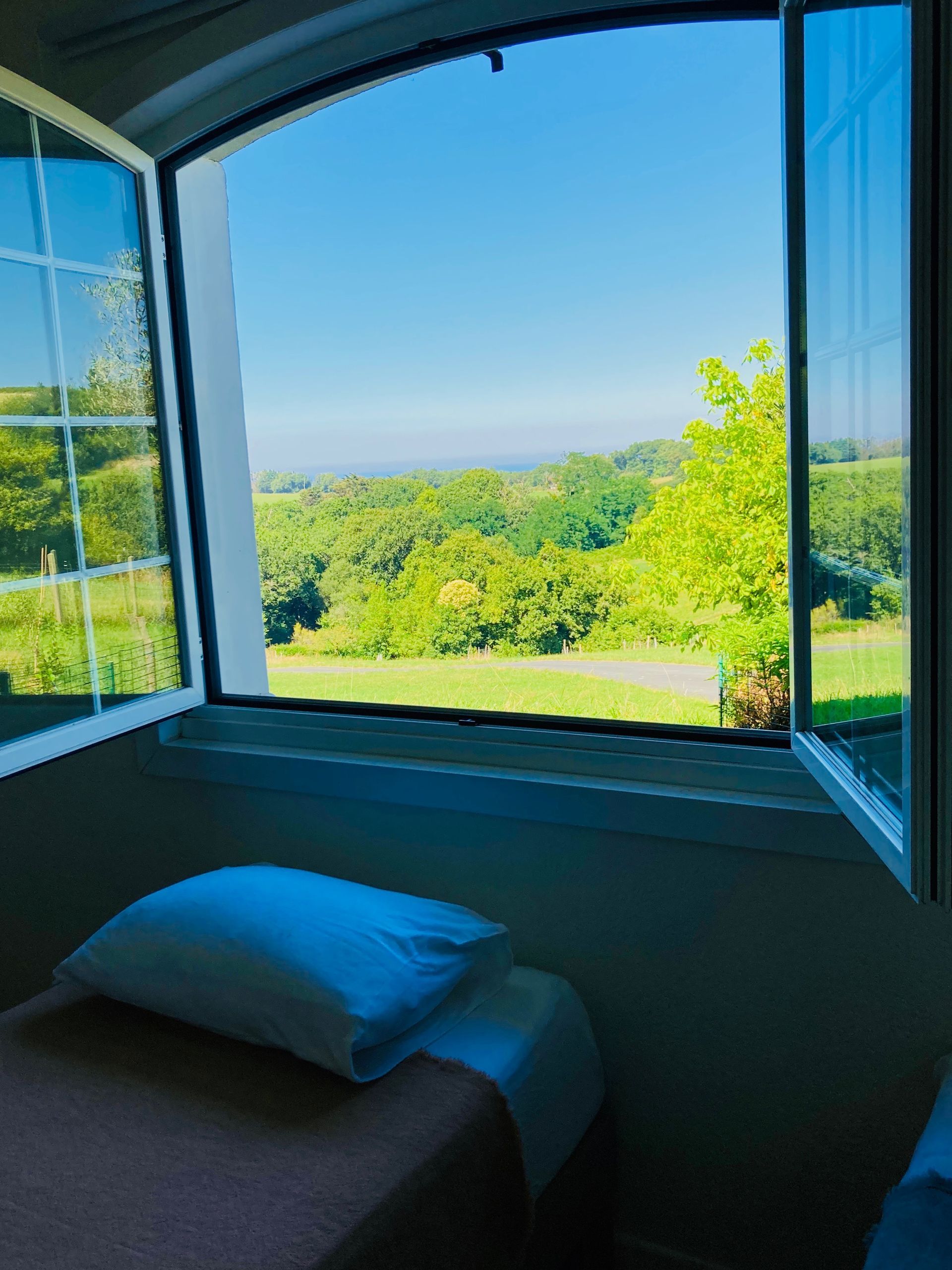 Open window with view of green hills, blue sky, and two beds with white pillows.