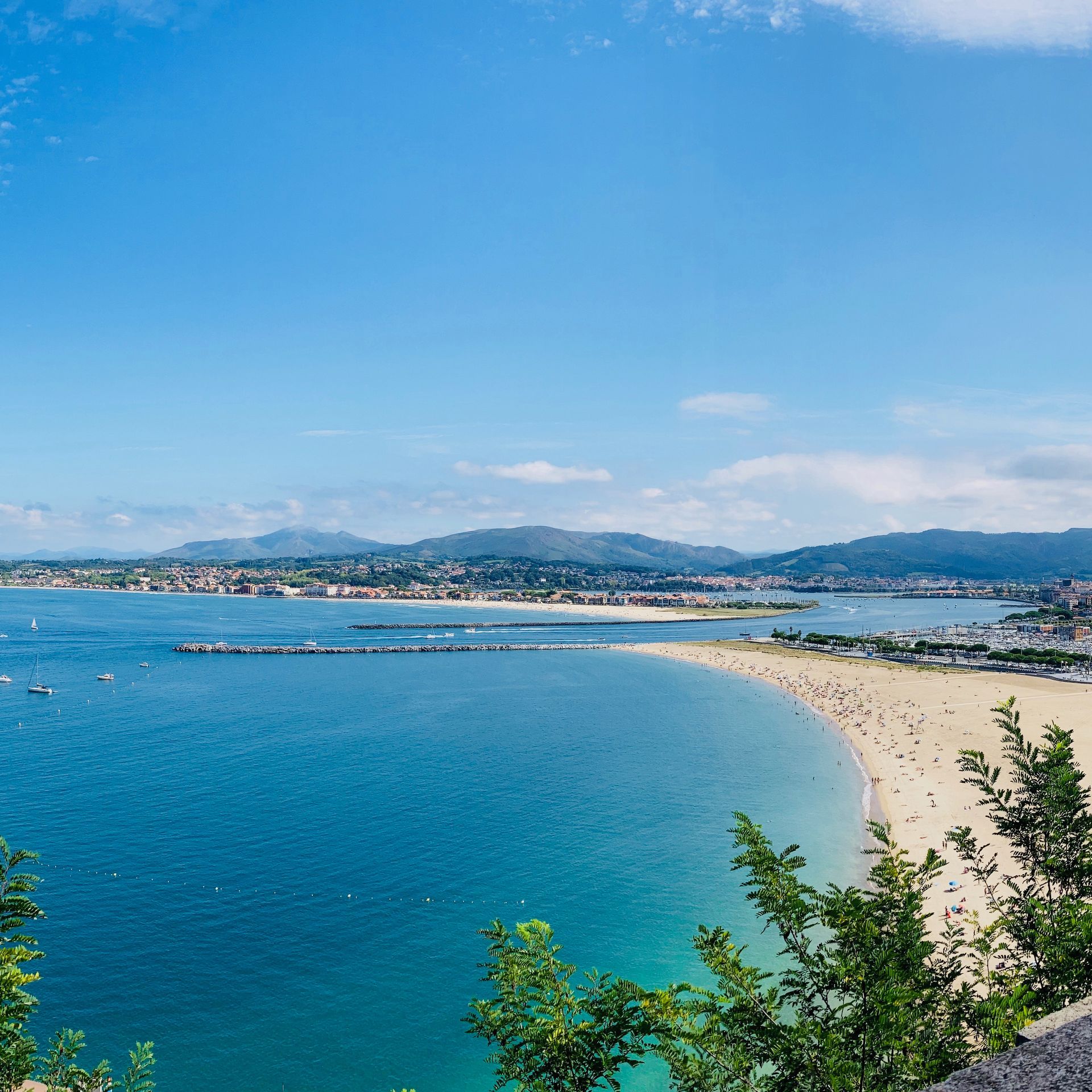 Scenic view of a sunny beach with blue water, a sandy shore, and distant buildings under a clear sky.