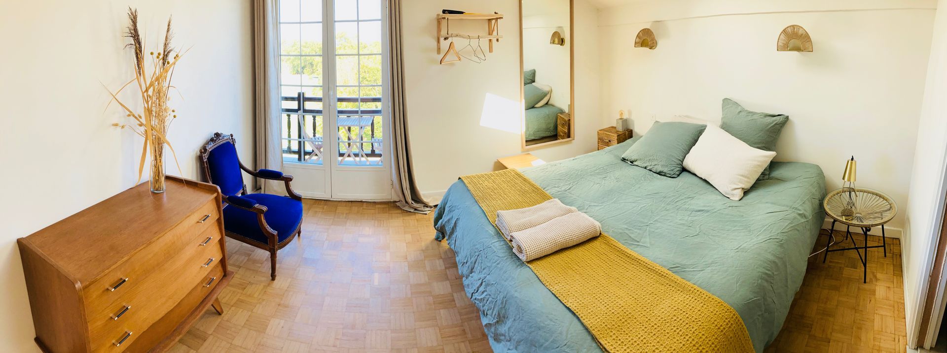 Bedroom with a large bed, dresser, and blue armchair, a balcony, and a large mirror.