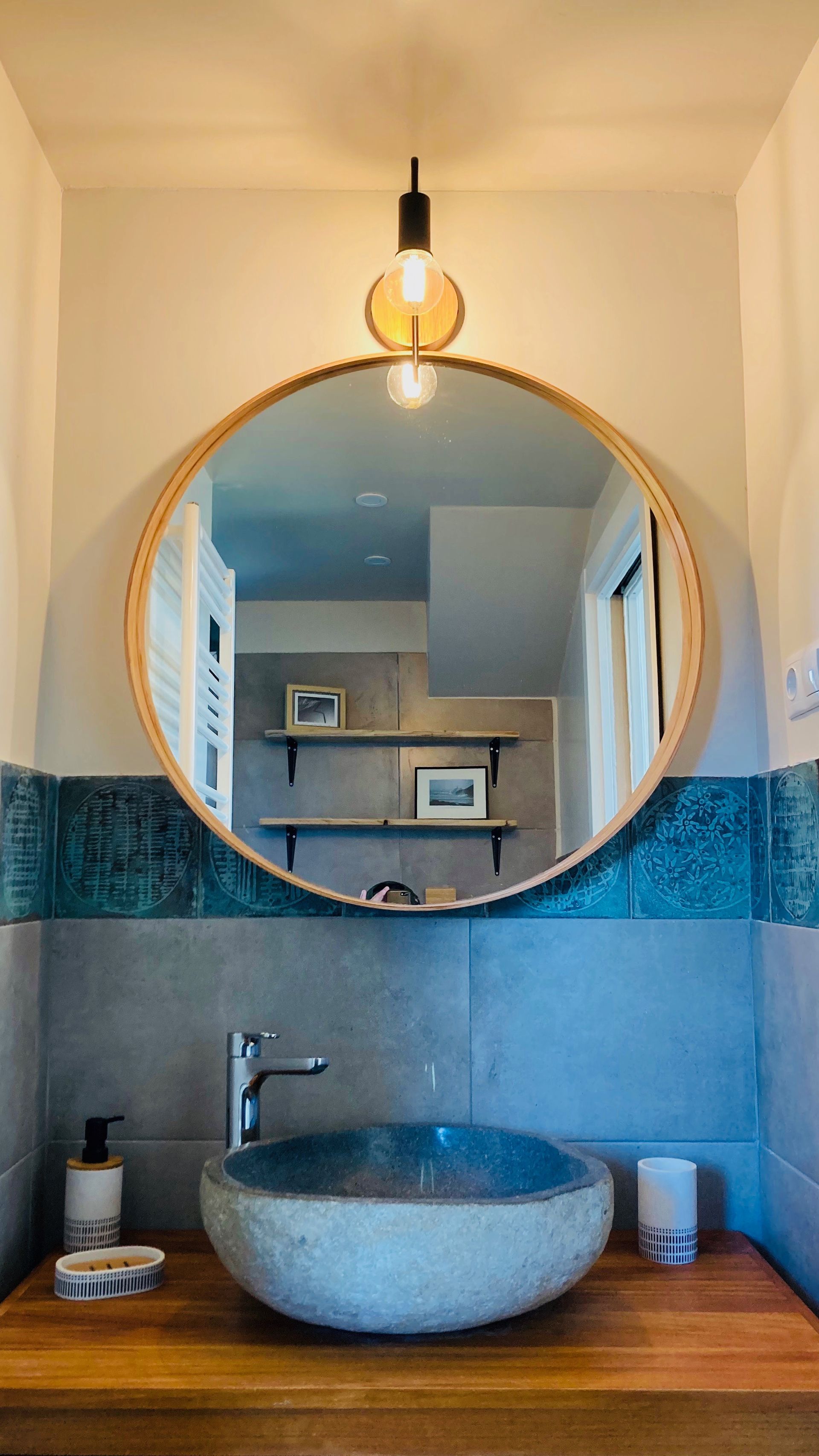Bathroom with round mirror, stone sink, wood counter, blue tile, and a hanging light.