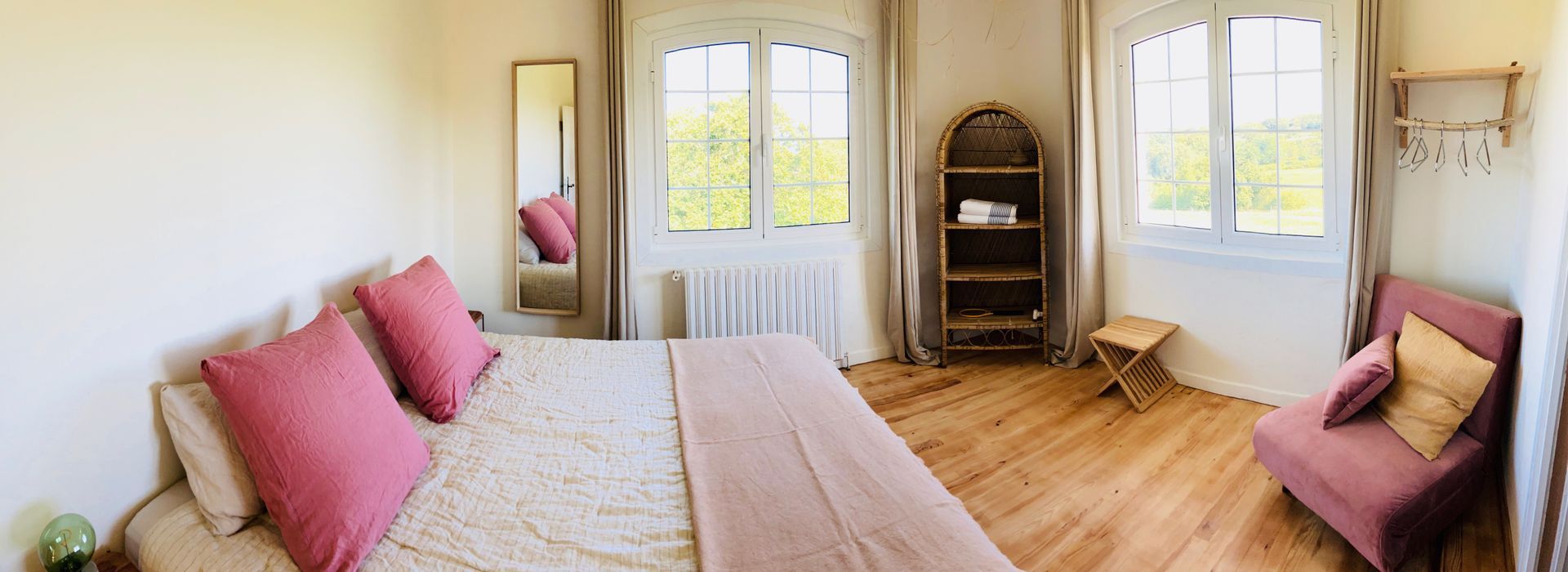 Bedroom with bed, mirror, window, small sofa, and wooden floor. Pink and cream tones.