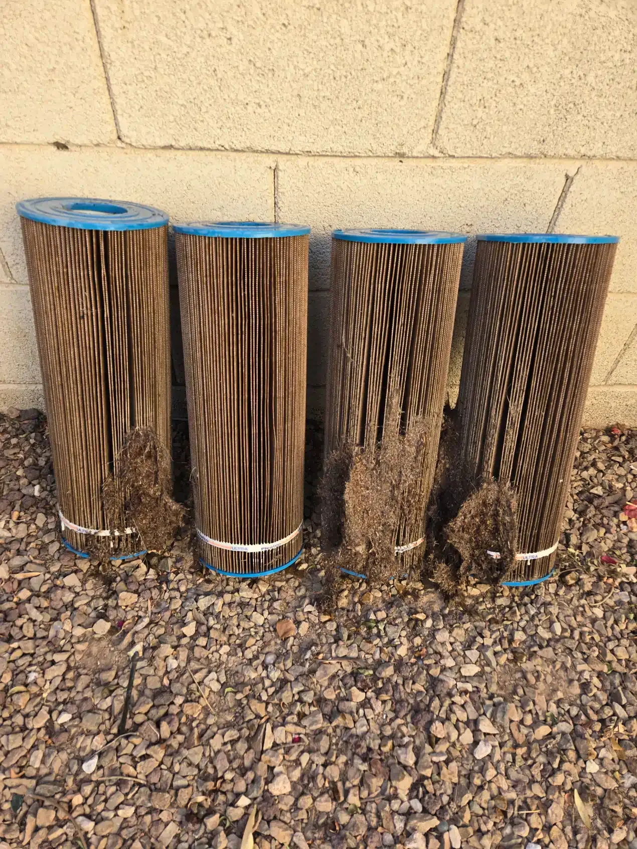 Four dirty filter cartridges are sitting on a pile of gravel in front of a brick wall.