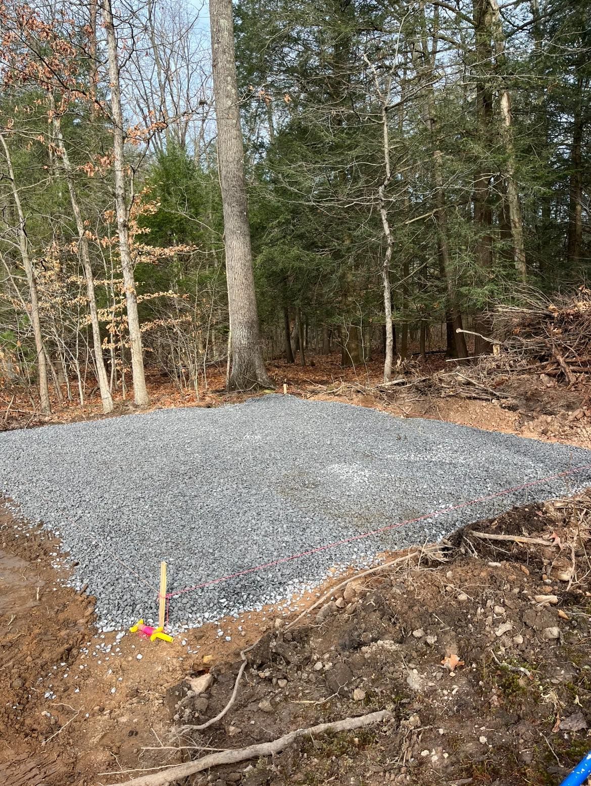 Gravel base construction in a wooded area. Gray gravel covers a cleared, rectangular area on uneven dirt. Trees in the background.