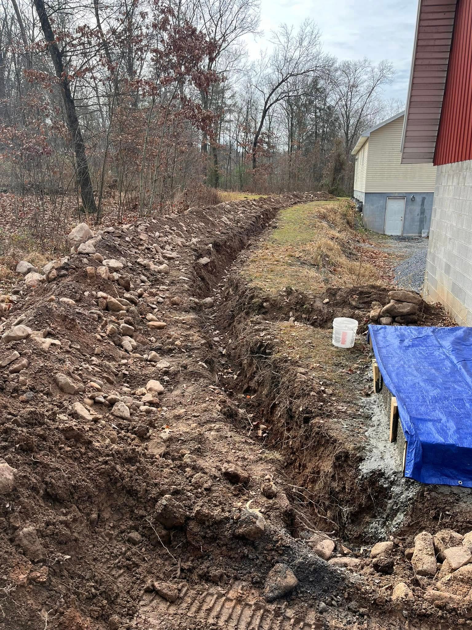 A trench dug in dirt next to a building, with trees in the background. A blue tarp and bucket are visible.