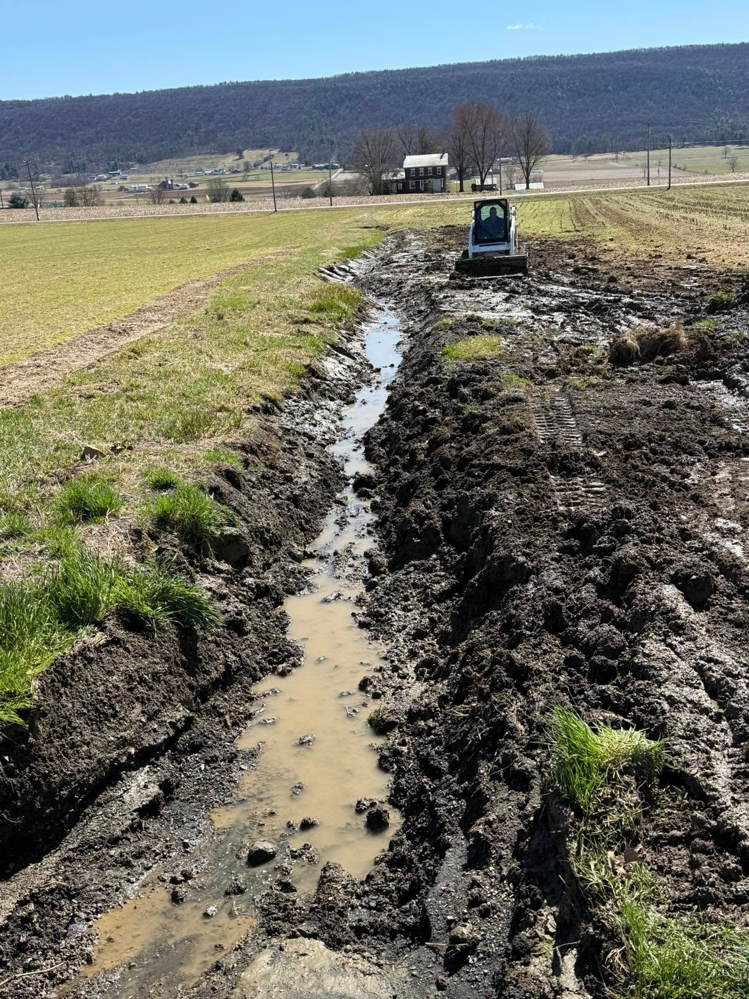 Mini excavator digging a water channel in muddy field, rolling hills in background.