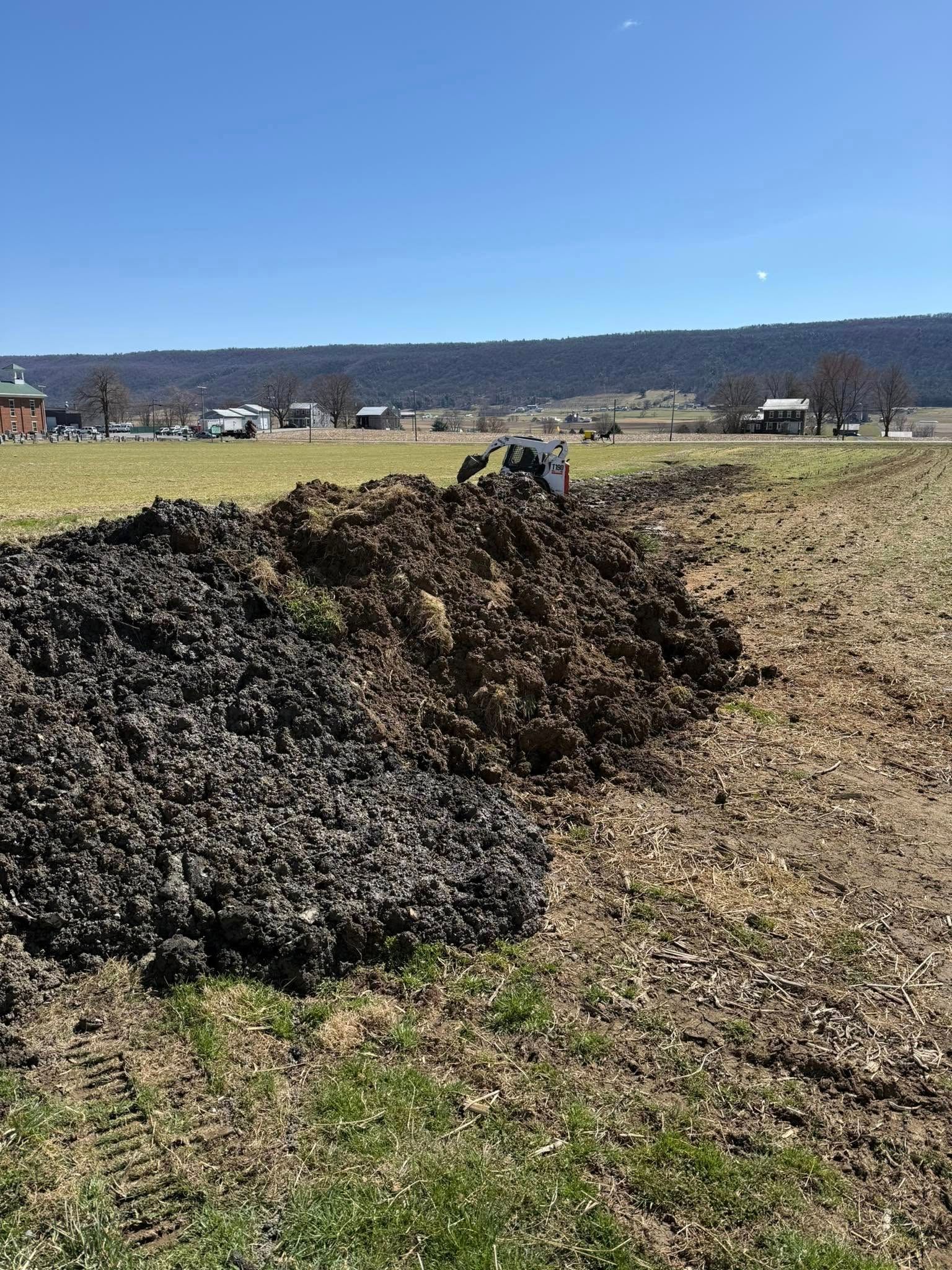 A large pile of dark soil sits next to a tilled field with a small tractor in the distance under a blue sky.