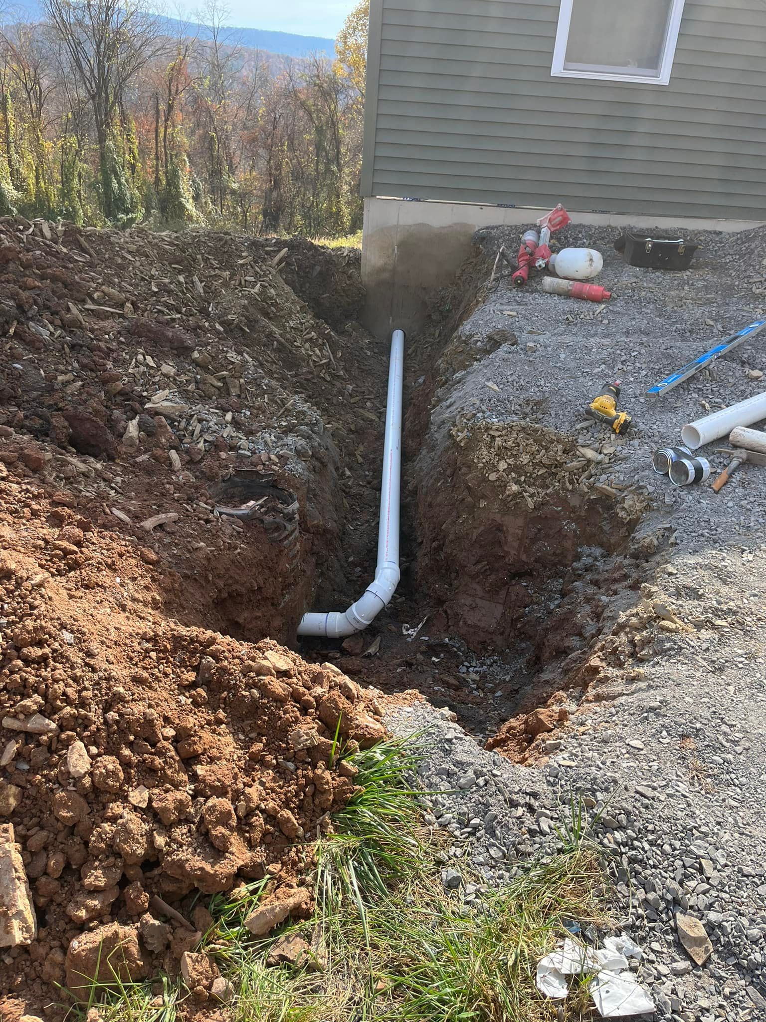 White PVC pipe in a trench, next to a building. Dirt and gravel surround.
