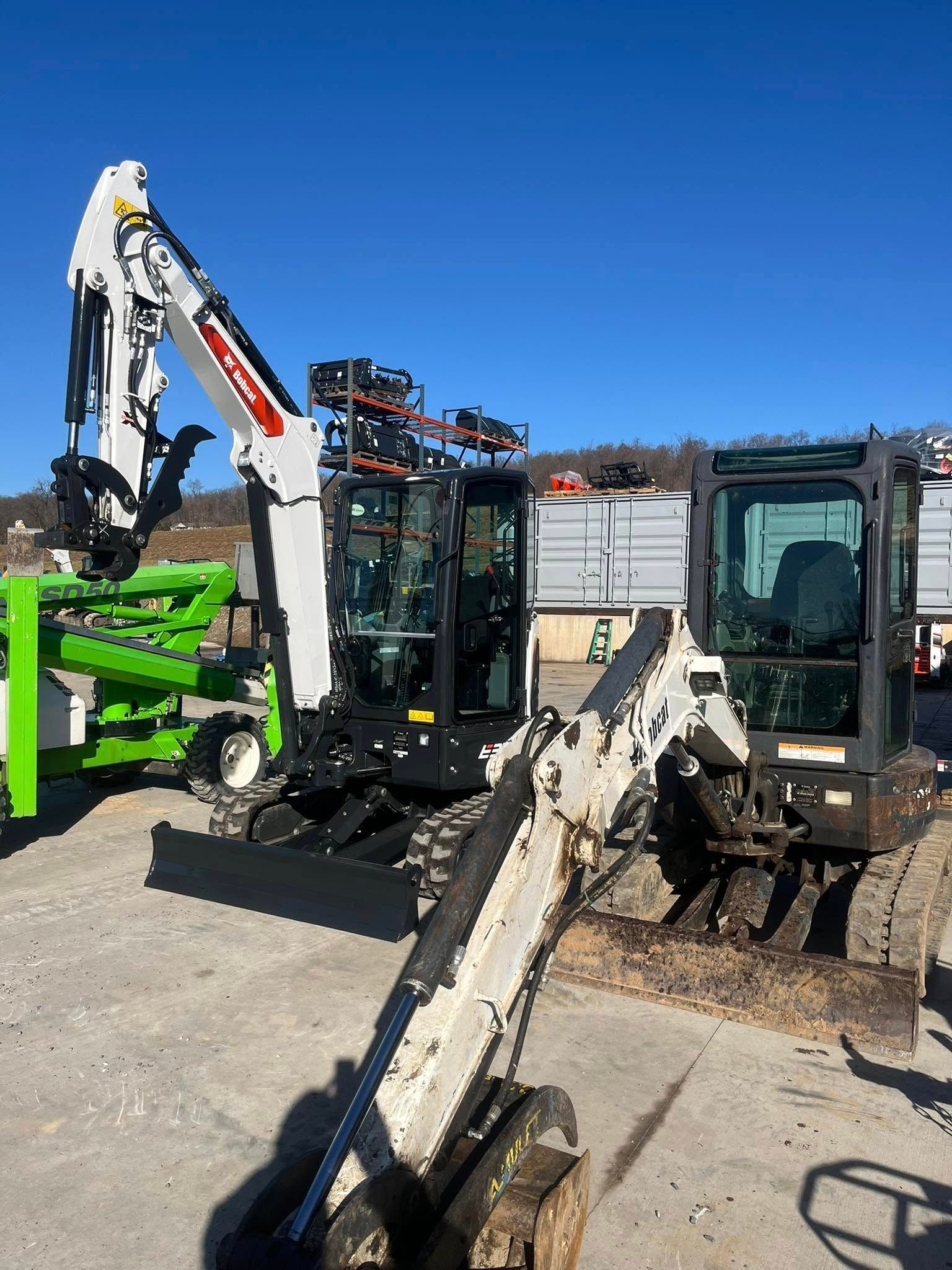 Two Bobcat excavators on a concrete surface, bright blue sky in the background.