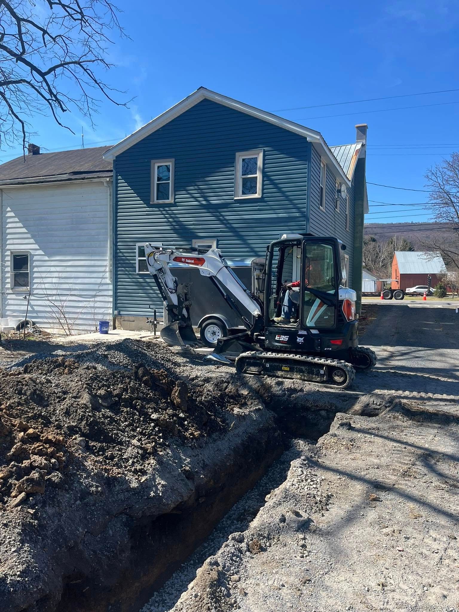 Excavator digging trench near blue and white houses on a sunny day.