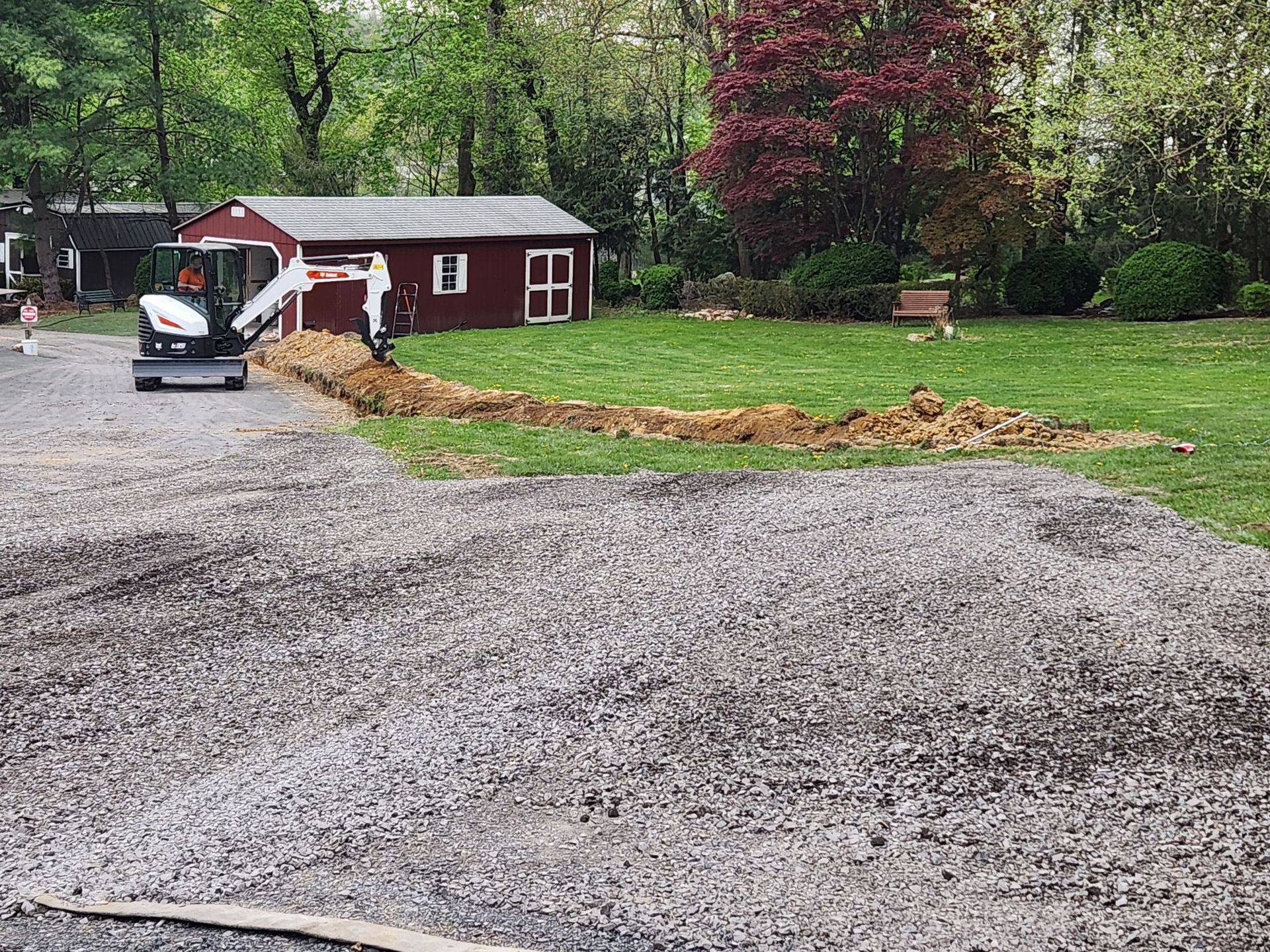 Bobcat excavator digging a trench on a grassy lawn next to a gravel driveway and a red shed.