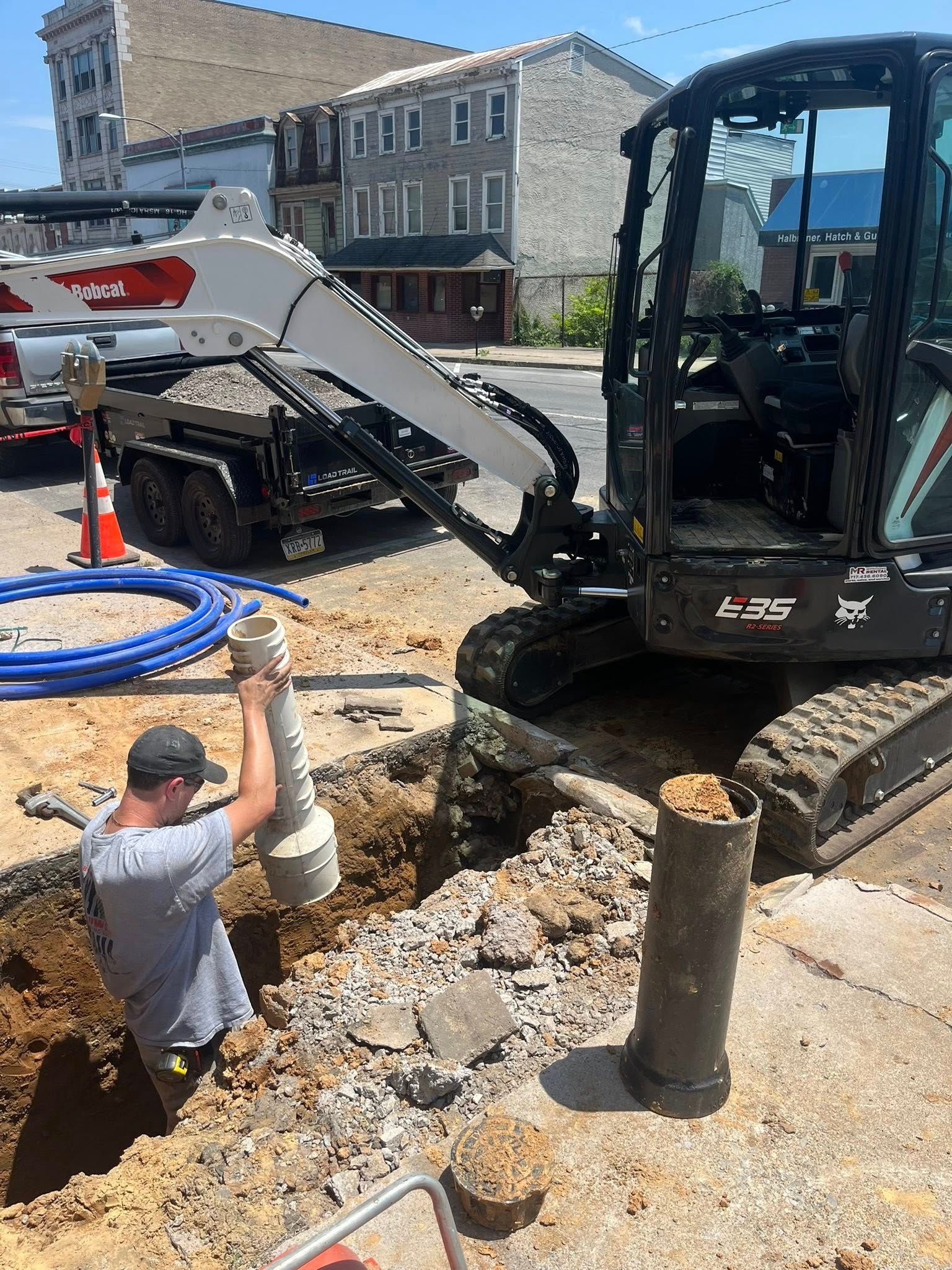 Construction worker installing a pipe in a street excavation, with an excavator and other equipment nearby.