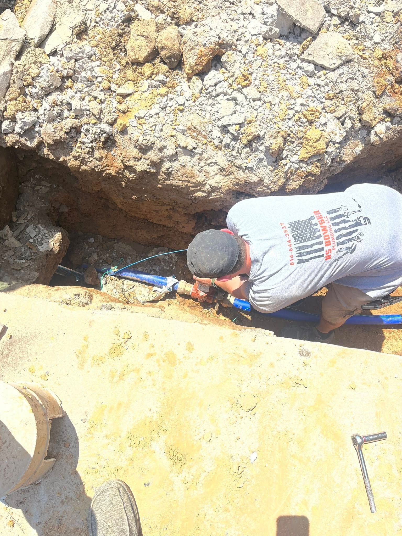Plumber repairing a blue pipe in a trench, water spraying. Sunny outdoor setting.