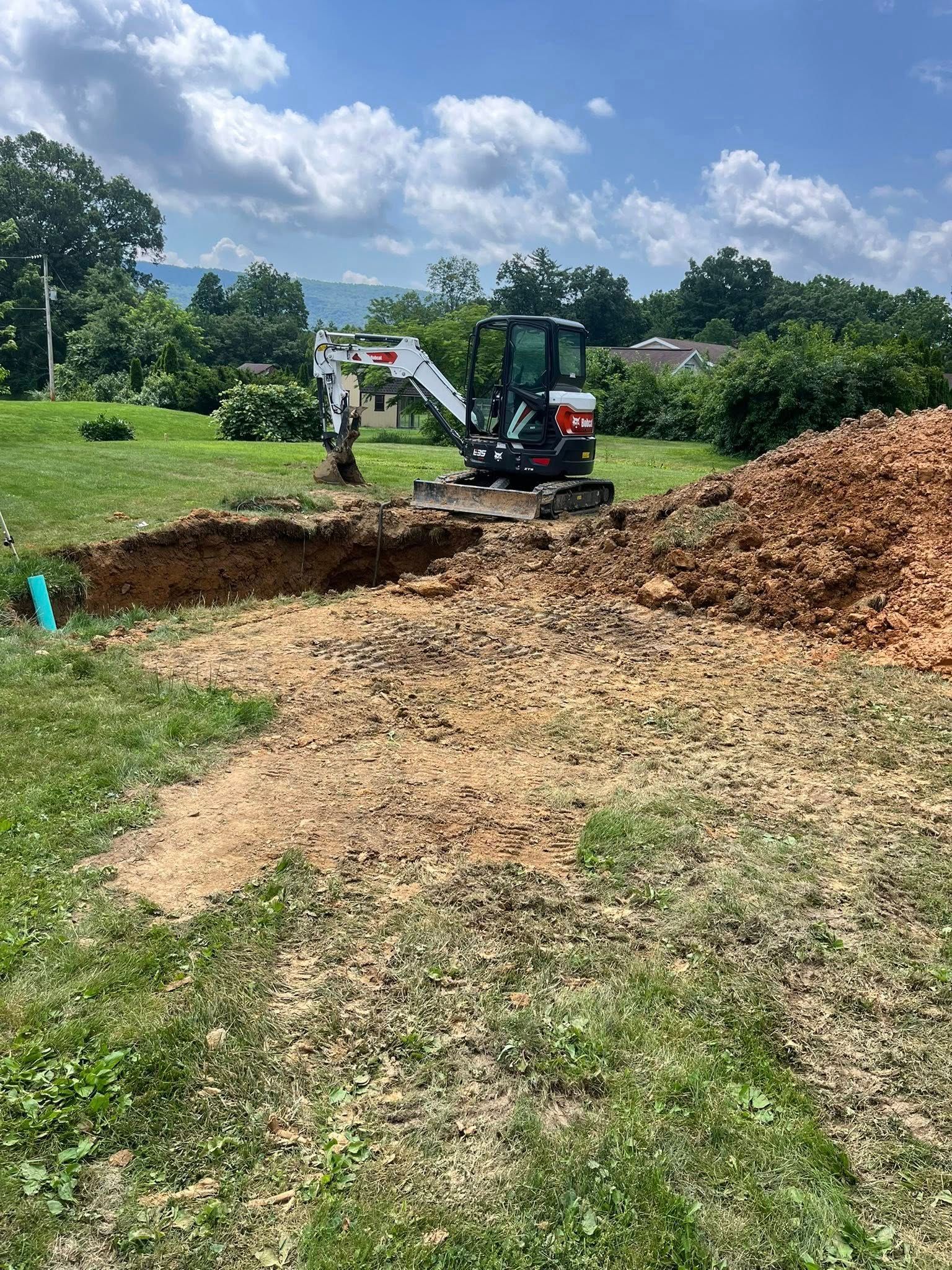 Bobcat excavator digging a trench in a grassy yard under a blue sky, with a pile of dirt nearby.