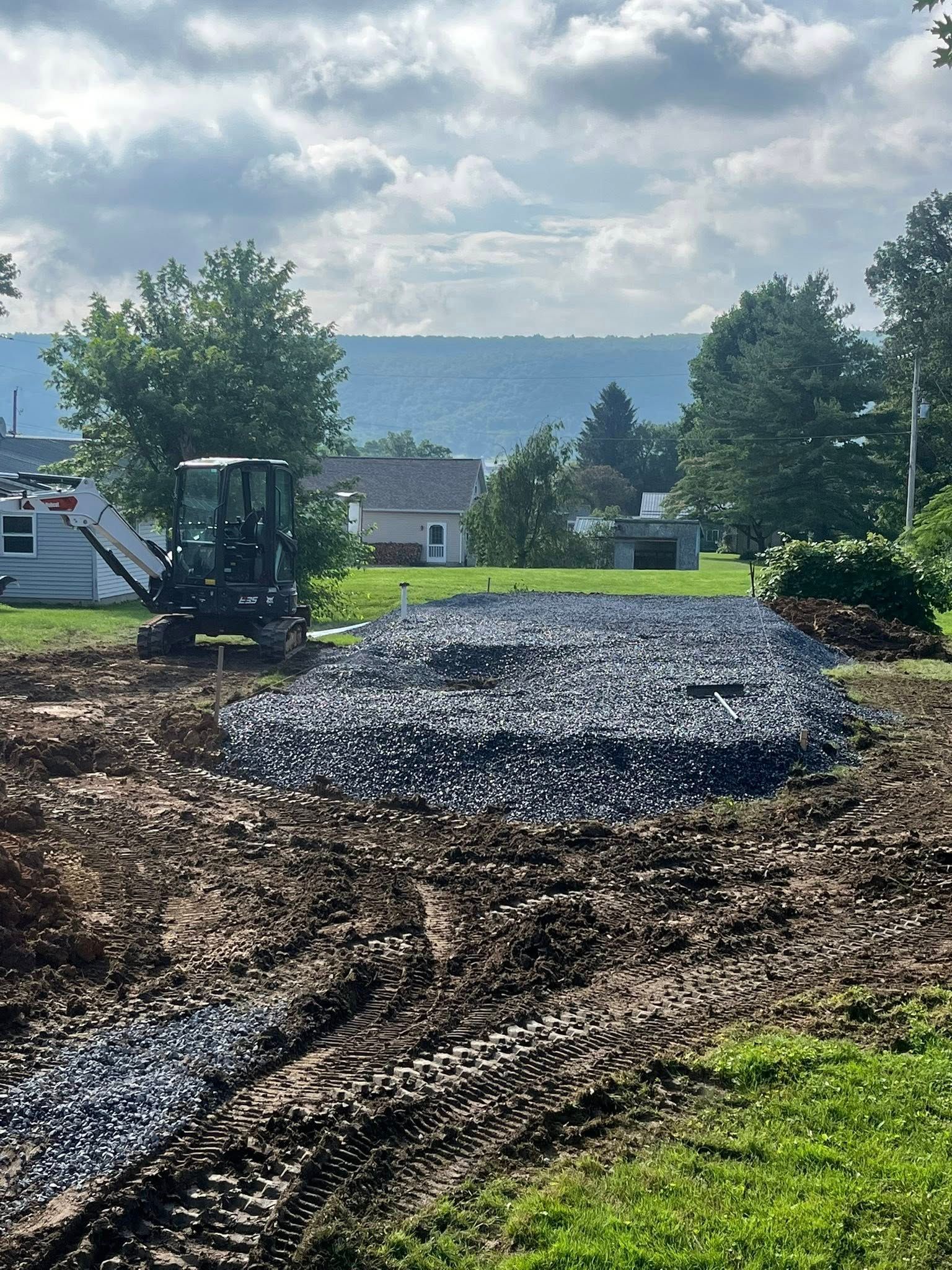 Gravel and an excavator on a dirt plot with a distant view of houses and hills under a cloudy sky.