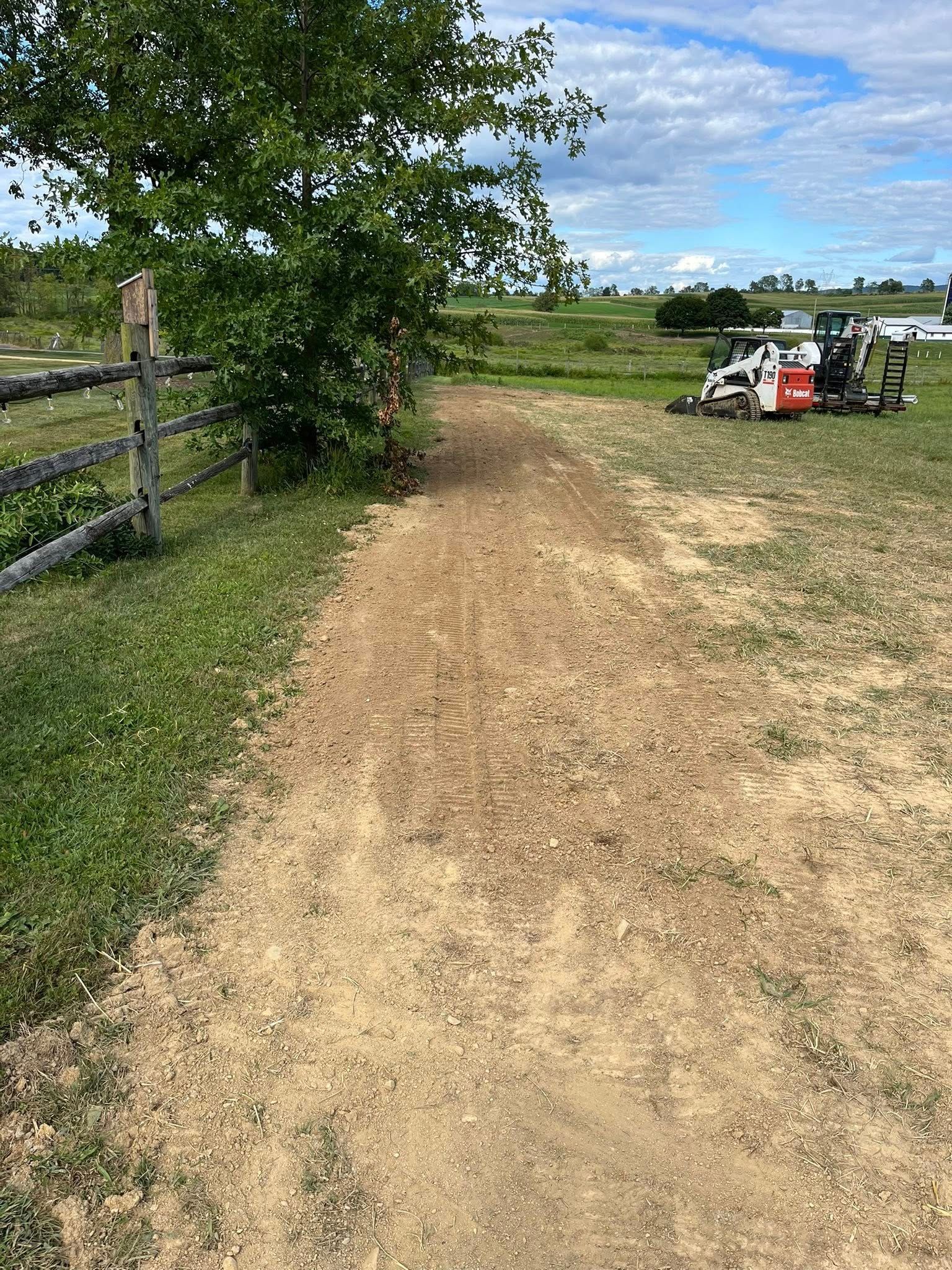 Dirt path leading through field next to a wooden fence and tree, with machinery in the distance.