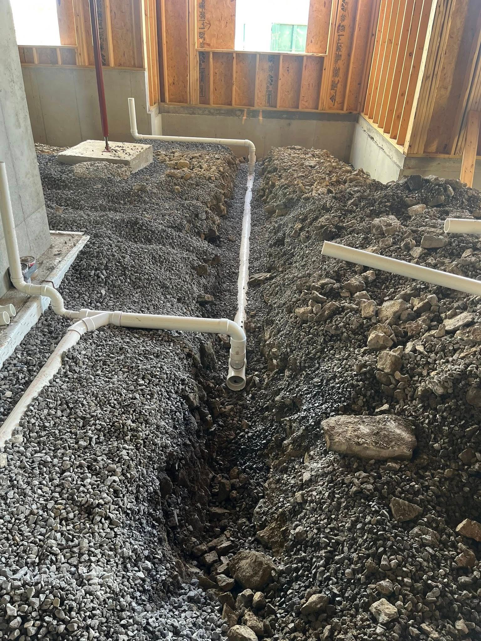 White PVC pipes run through gravel base inside unfinished building. Wooden frame visible.