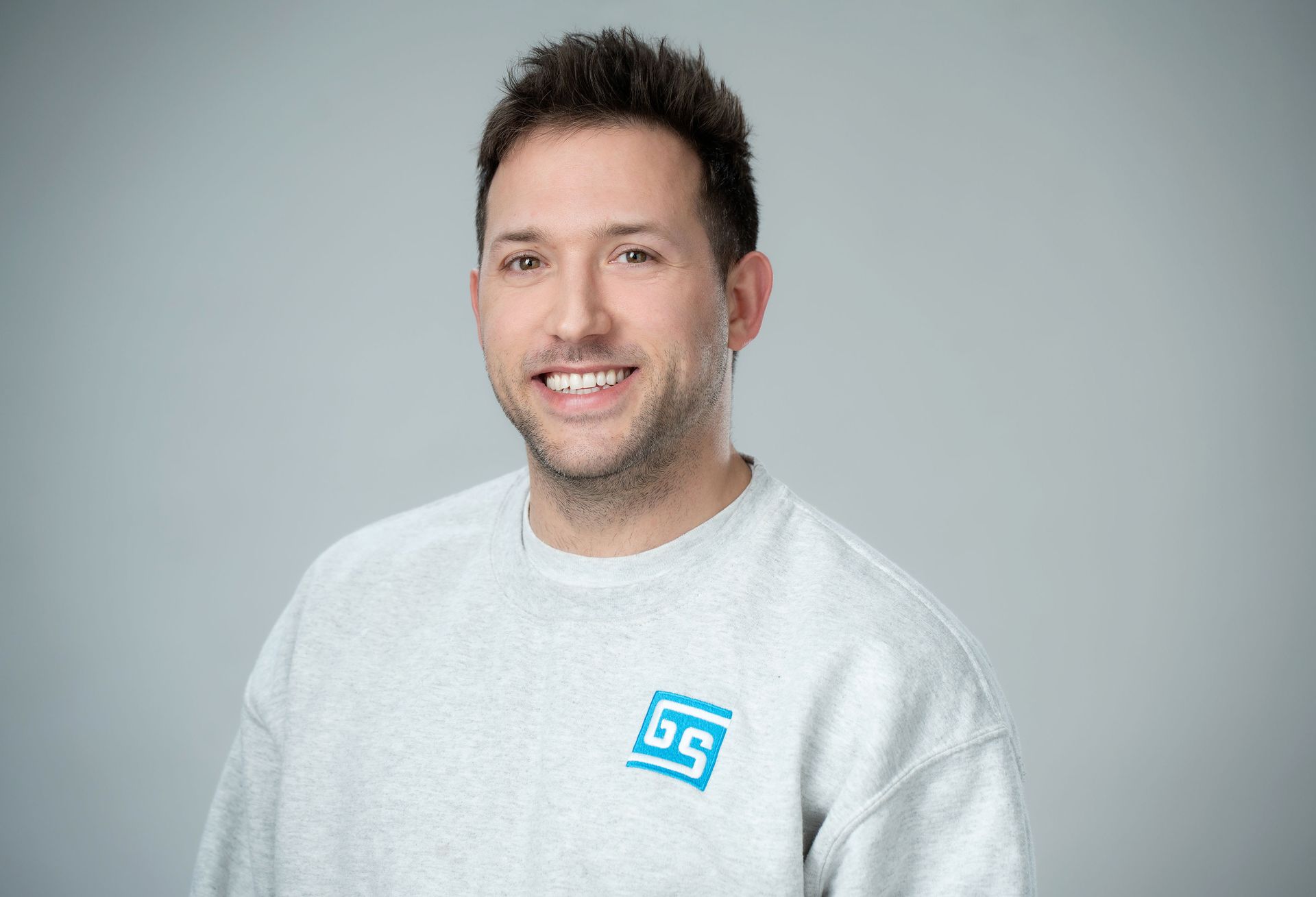 A man wearing a grey sweatshirt with a blue logo on the front is smiling for the camera.