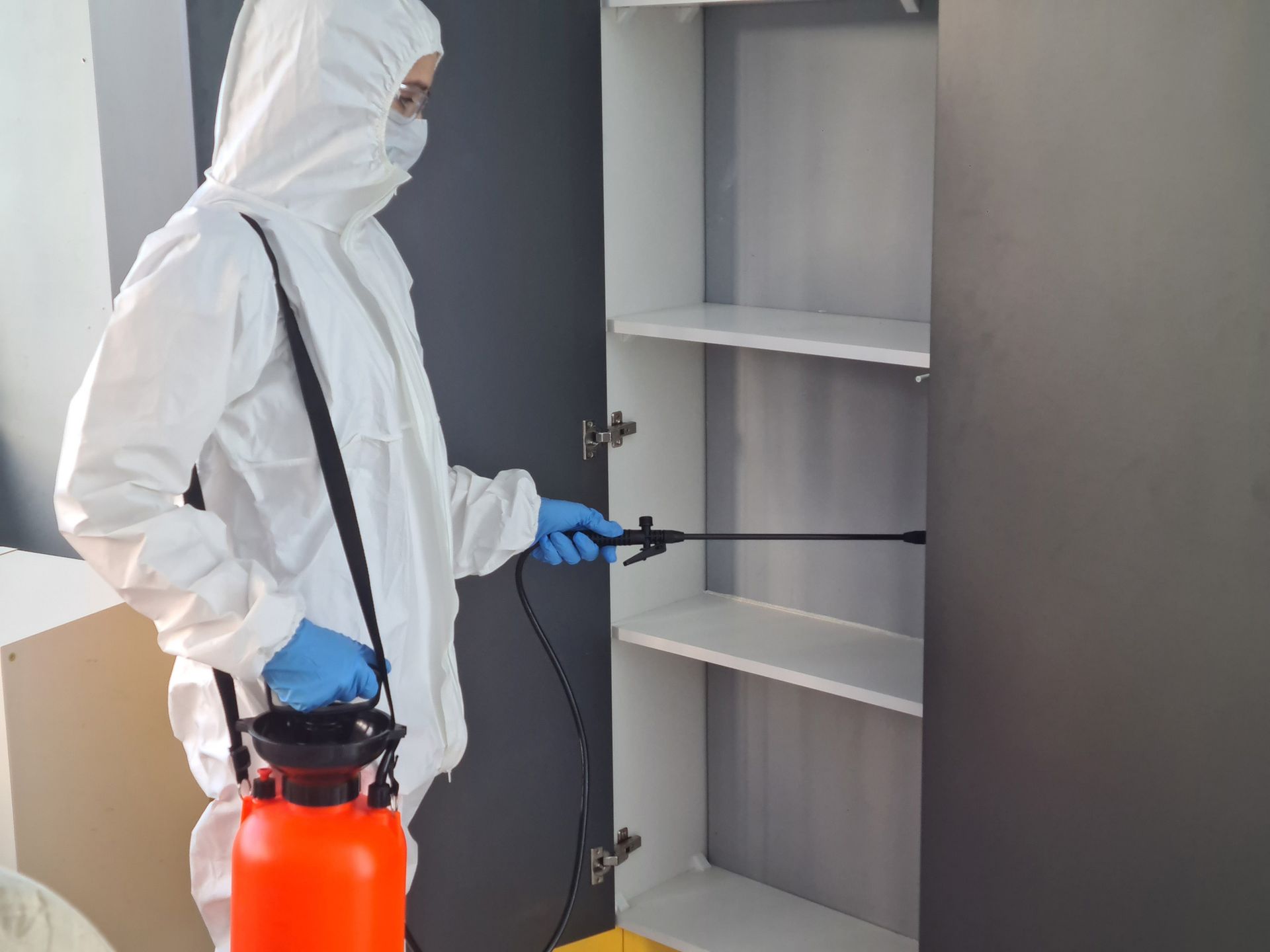 Technician in protective gear sprays inside cabinet during pest control treatment.