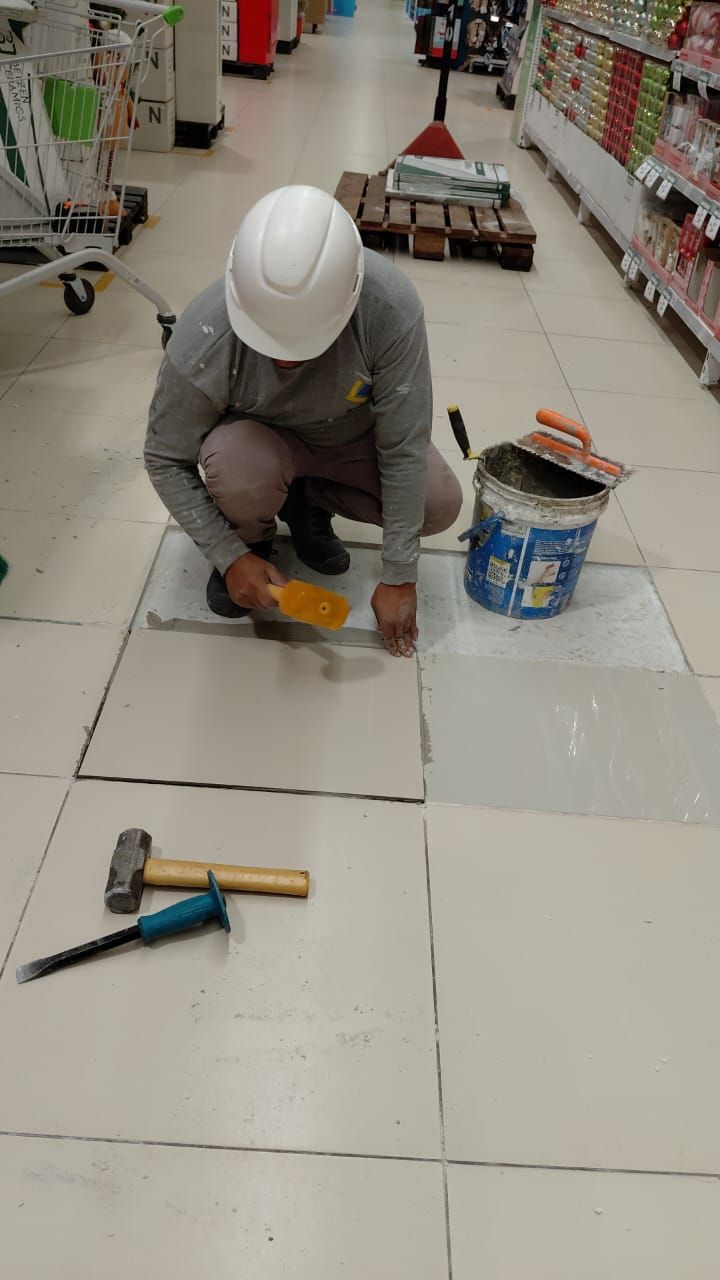 Un trabajador con casco blanco instala baldosas en el suelo de una tienda, utilizando una llana y un cubo de adhesivo que tiene cerca.