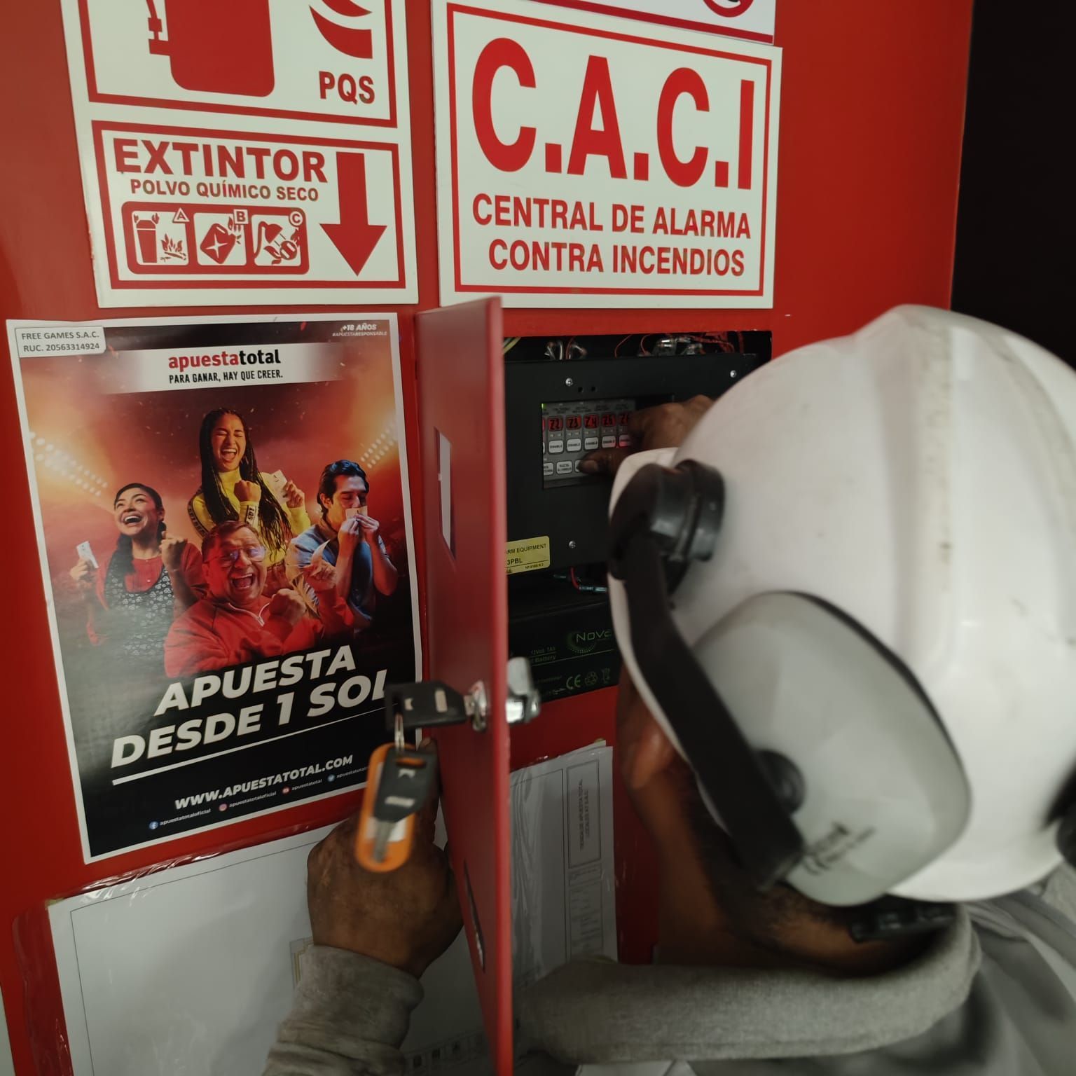 Un trabajador con casco y orejeras realiza el mantenimiento de un panel de control rojo de la alarma contra incendios, situado junto a un anuncio de juegos de azar.