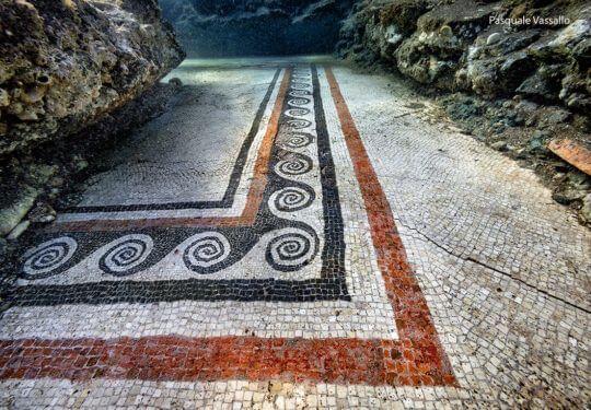 Underwater view of a mosaic floor with black, red, and white patterns, between rock walls.
