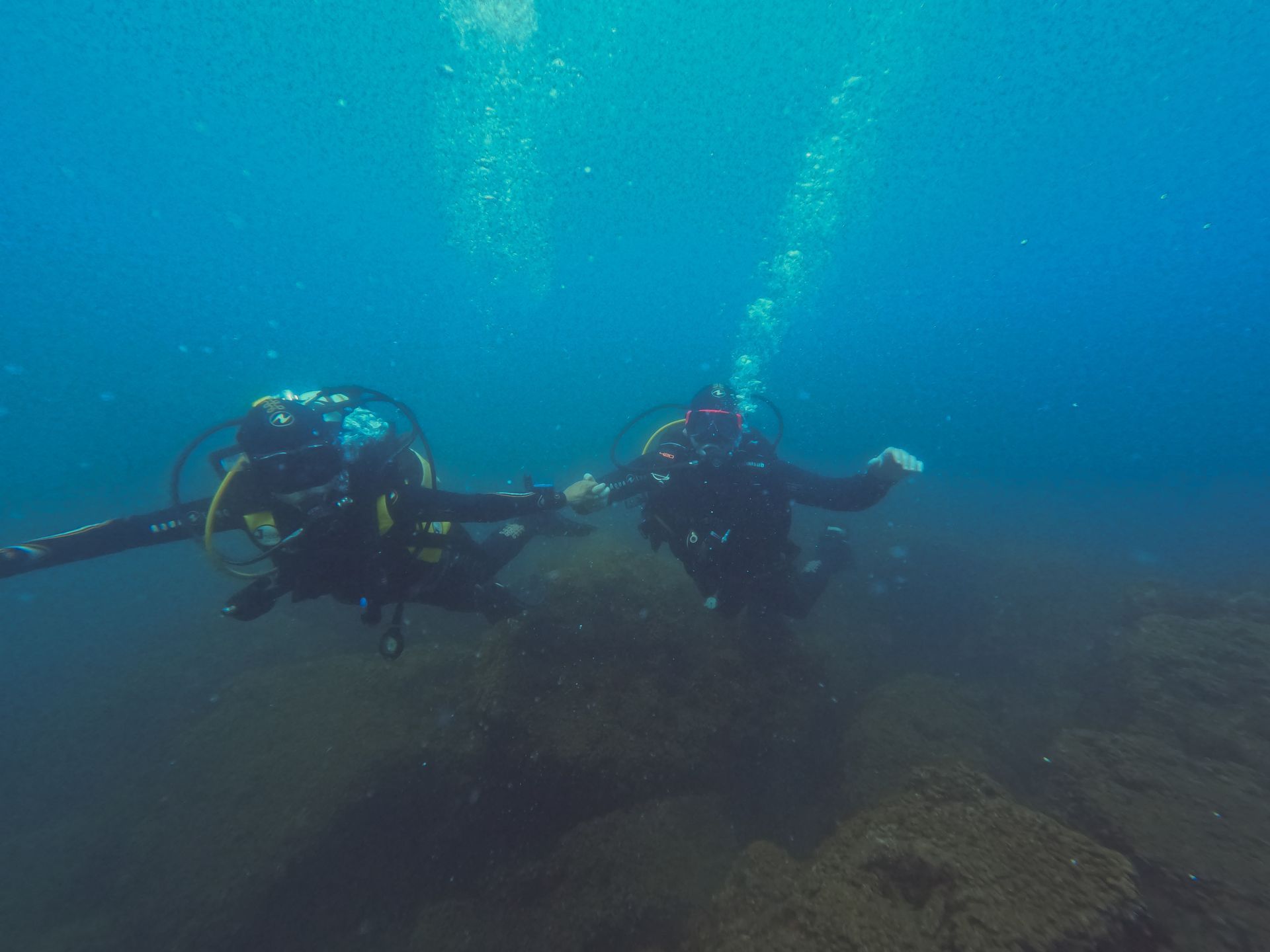 Two scuba divers holding hands underwater, blue water, dark rocks in background.