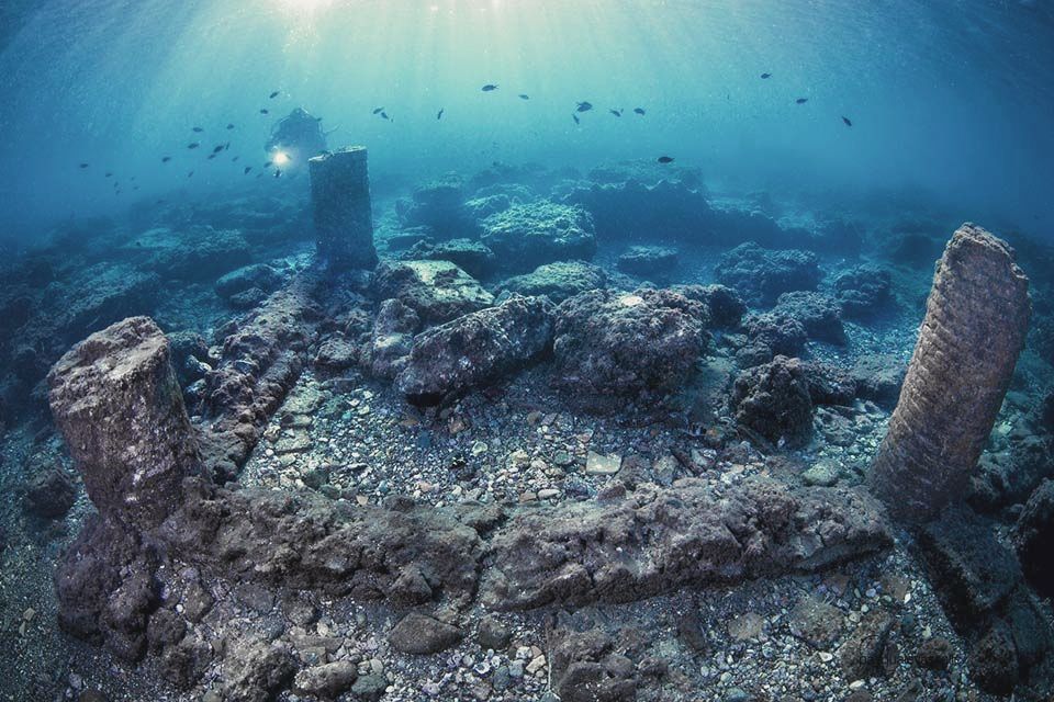 Underwater view of ancient ruins, including columns and a diver exploring a seabed.