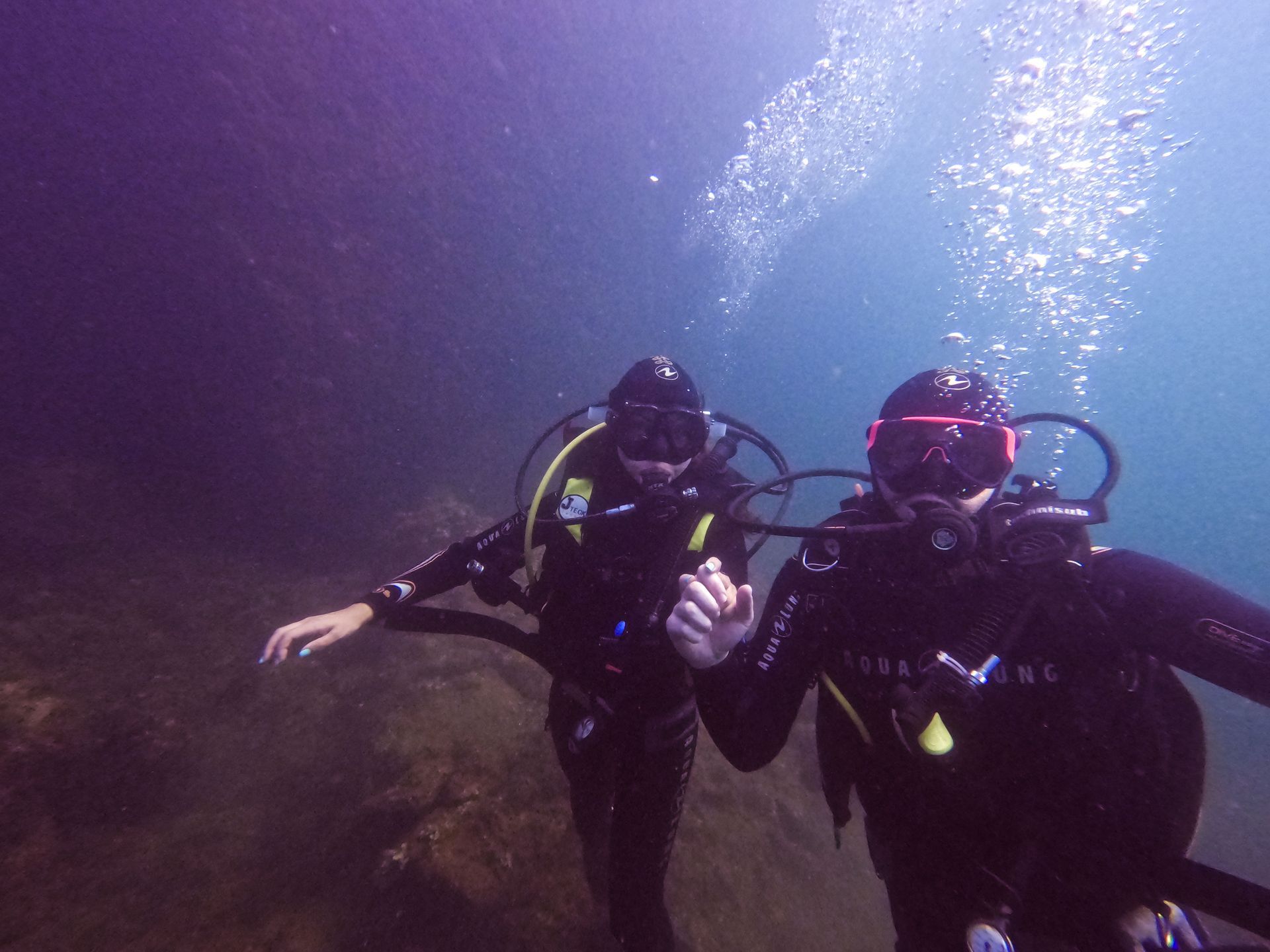 Two scuba divers explore underwater, near a rock formation. Bubbles rise; one diver gestures.