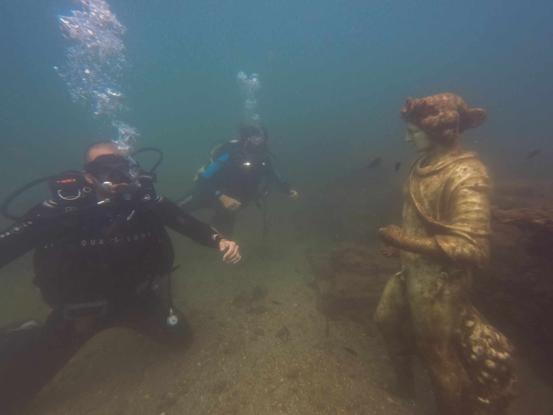 Divers explore an underwater statue in murky water.