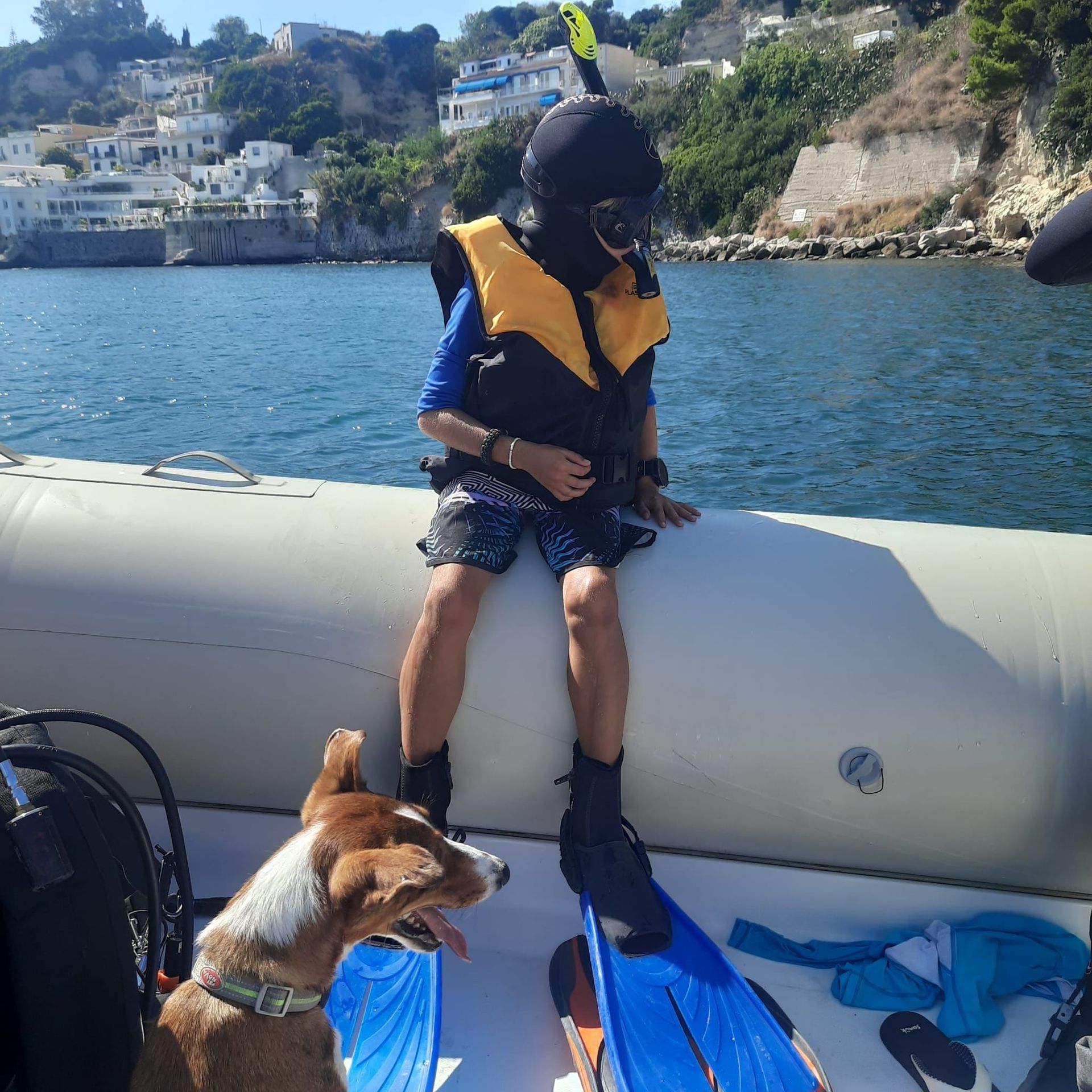 Person in dive gear on inflatable boat with a dog, ready to snorkel; sea and buildings in background.