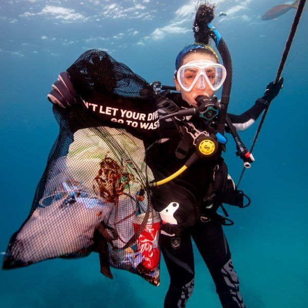 Scuba diver collecting trash underwater; holding a net with debris, blue water. 