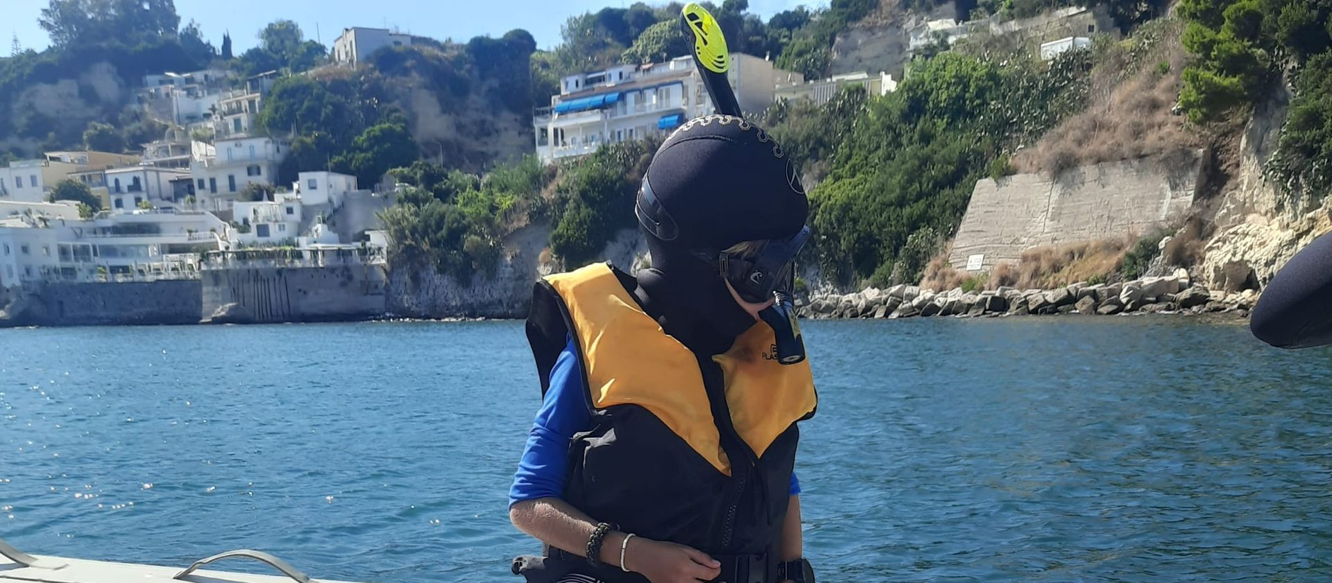 Person in scuba gear on a boat, facing forward. Water and white buildings in the background. Sunny day.