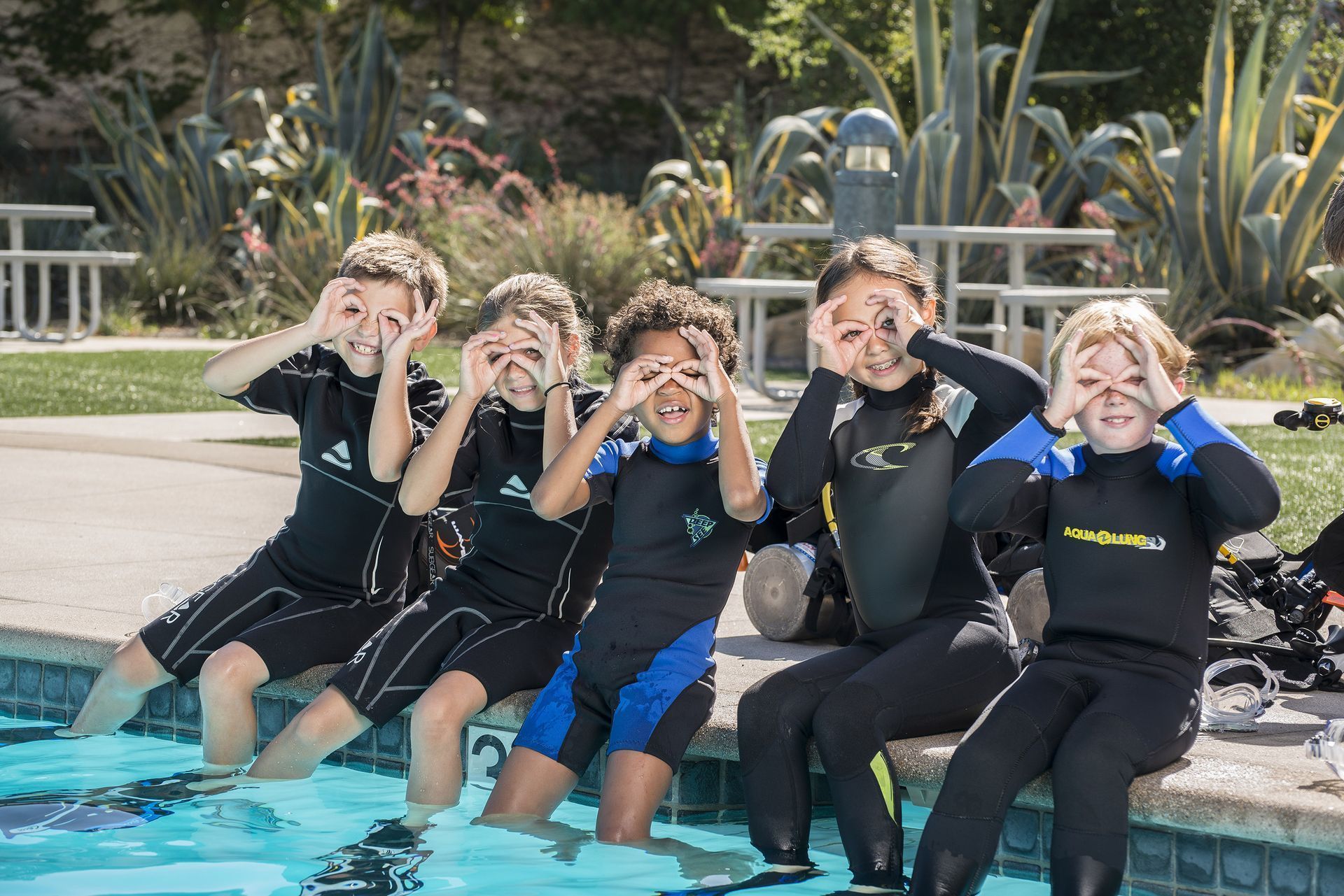Five children in wetsuits by a pool, making eye circles with their fingers.