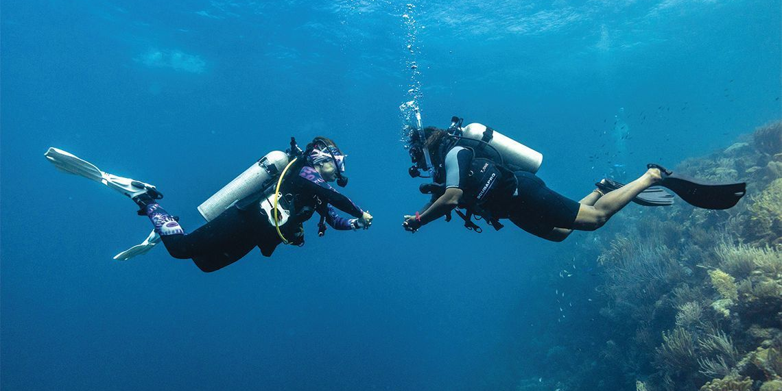 Two scuba divers in blue water, facing each other. Bubbles rise above one of the divers near a coral reef.