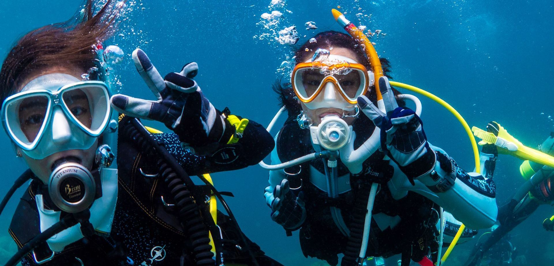 Two scuba divers underwater giving the peace sign. They wear masks, tanks, and wetsuits, surrounded by bubbles.