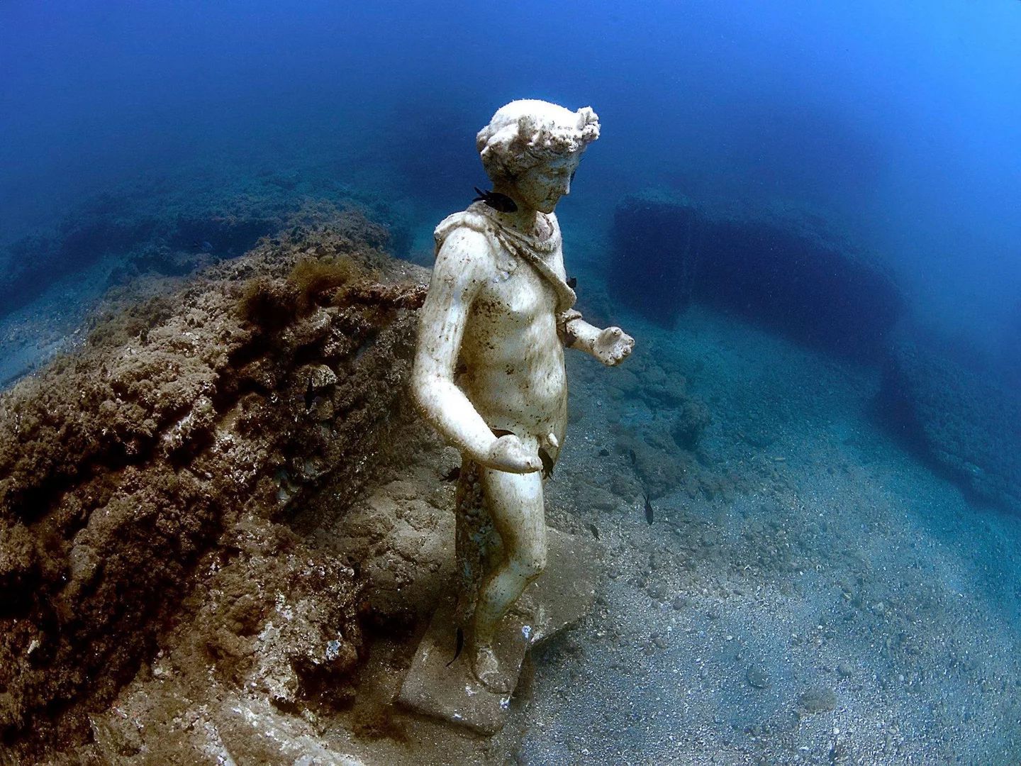 Underwater statue of a classical figure, partially covered in marine growth, on a seabed with clear blue water.