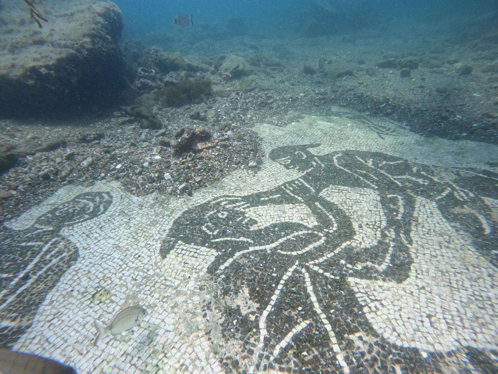Underwater mosaic with black and white tiles depicting figures in a sandy sea bed.