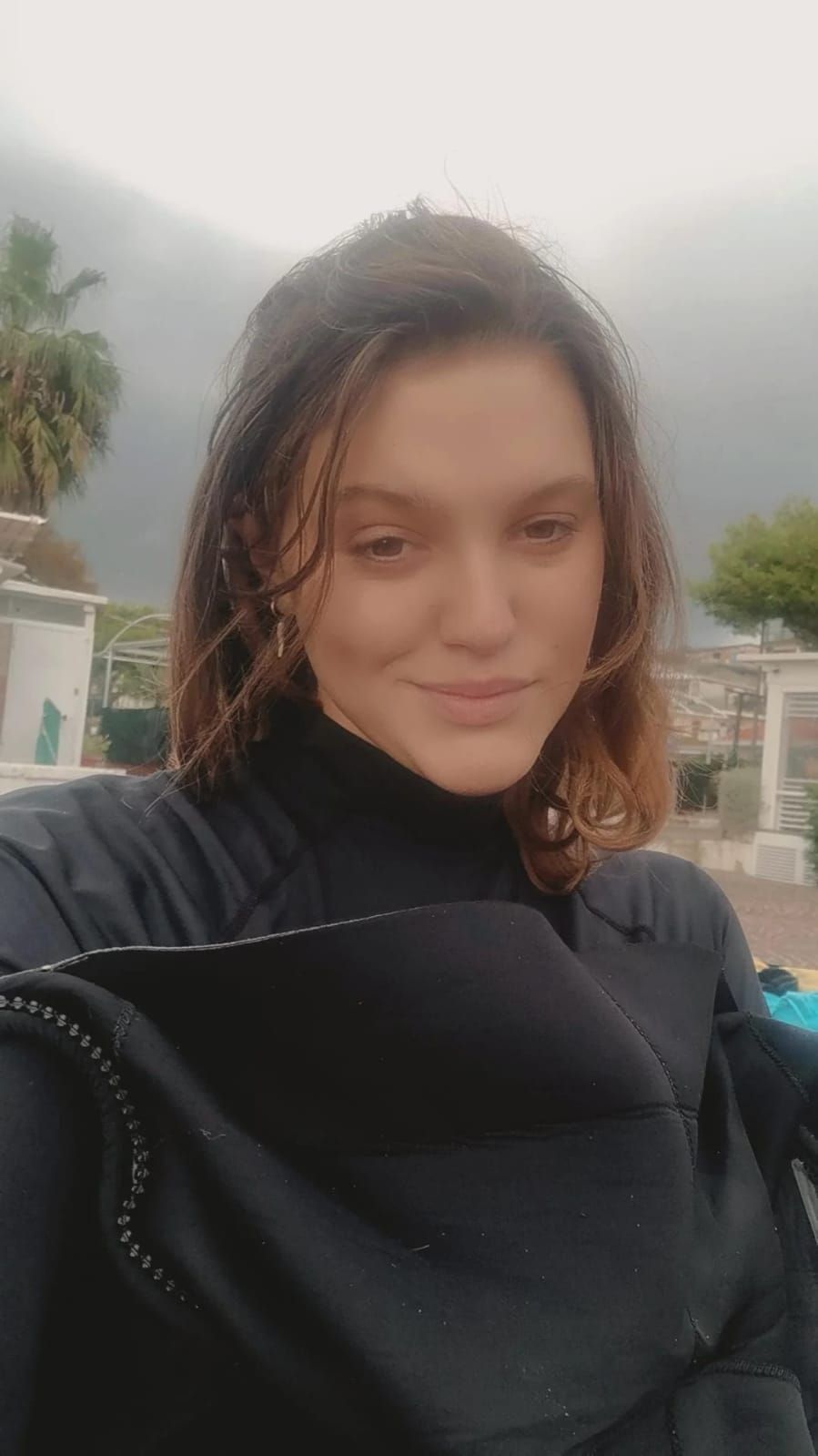 Woman in a wetsuit, wet hair, looks at the camera. Outdoor setting, cloudy day.